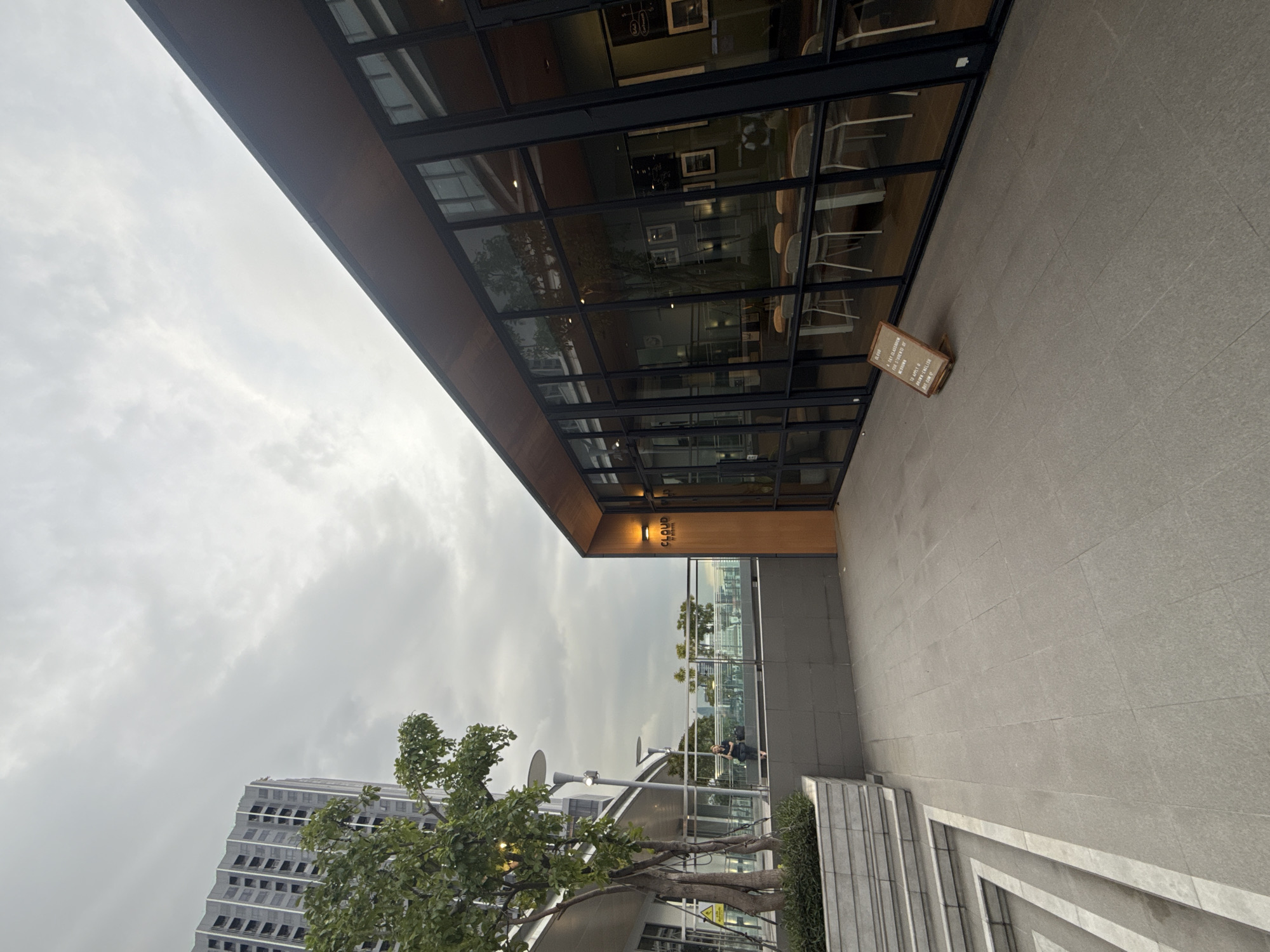 The image shows an exterior view of a modern cafe or restaurant named "CLOUD" located in Bangkok, Thailand. The cafe features large glass windows and a wooden facade, with an outdoor seating area paved with large gray tiles. A small, A-frame sign with text is placed on the ground in front of the cafe. To the left, a modern tall building stands against a cloudy sky. A tree with green foliage is planted in a planter box, and a person is seated on a ledge in the background. The overall scene suggests a contemporary urban environment.