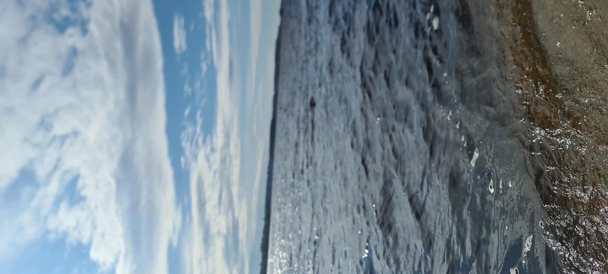 The image captures a natural landscape scene in Ikšķile, Latvia, under a clear daytime sky. The composition is divided into three main horizontal sections when viewed correctly.
The top third of the image features a bright, light blue sky adorned with scattered white clouds. Some clouds are thin and wispy, while others are larger and more defined, suggesting fair weather.
A dark, thin horizontal line separates the sky from the water, representing a distant horizon or a low-lying landmass, possibly a forested bank.
The lower two-thirds of the image are dominated by a body of water. The middle section of the water appears relatively calm with gentle ripples, reflecting the sky and suggesting depth. However, the immediate foreground, particularly towards the right, shows highly turbulent water. This area is characterized by splashes, foam, and individual droplets sparkling in the light, indicating active movement such as a strong current, waves breaking, or water flowing over an unseen feature. The water in this turbulent section also displays varying shades, from deep blues to lighter, almost greenish-brown tones, possibly revealing the riverbed or sediment.
The scene conveys a sense of natural beauty and the dynamic interplay between the calm expanse and active movement of water. No people, animals, or man-made objects are visible, and there is no discernible text. The bright lighting indicates it is daytime, likely mid-morning or afternoon.