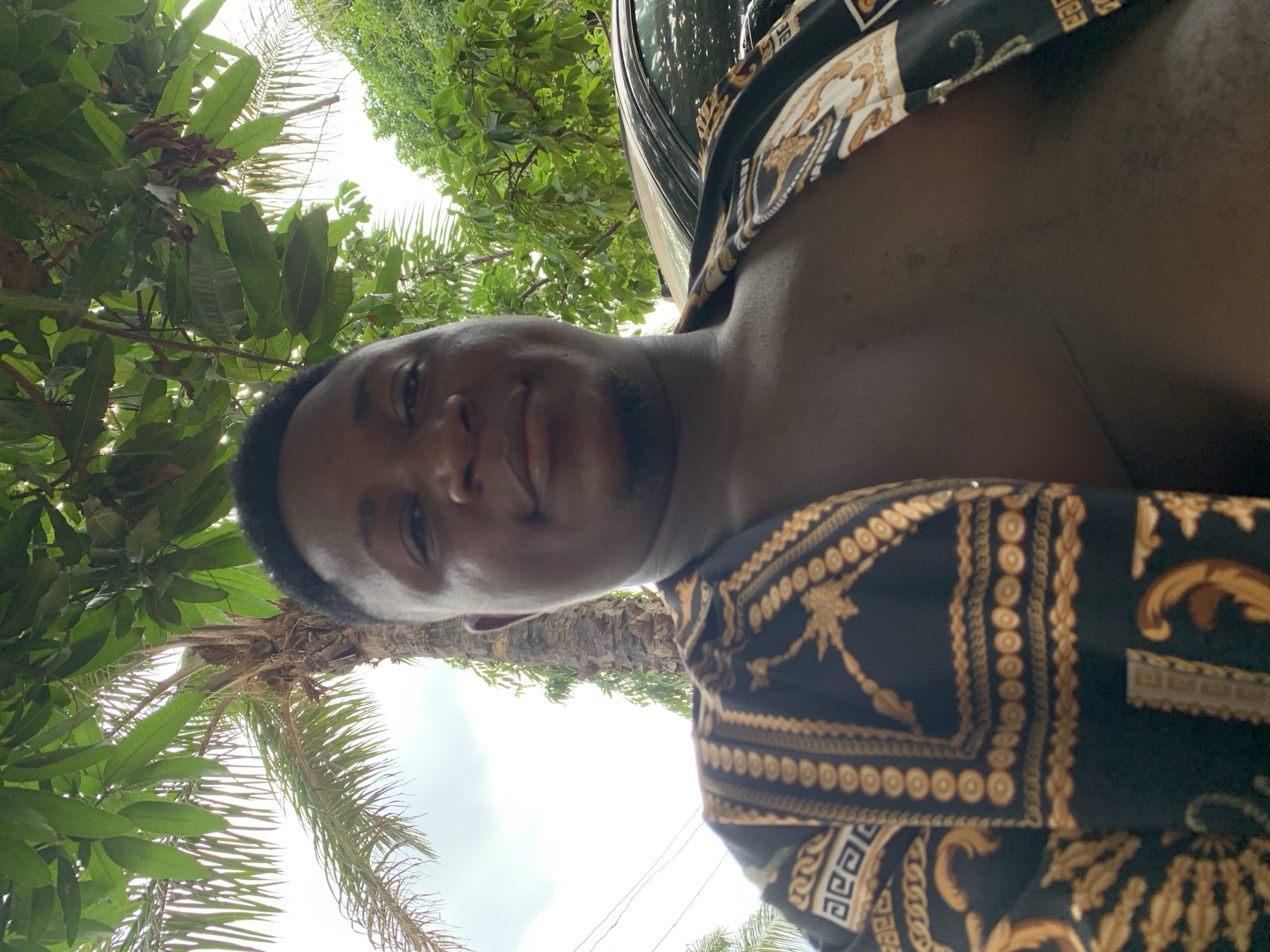 A low-angle selfie shows a dark-skinned man with closely cropped hair, wearing a black shirt with gold decorative patterns. The background includes a palm tree and other trees, suggesting a location in Akwanga, Nigeria. A car is visible partially at the top of the frame. The sky is visible through the trees.