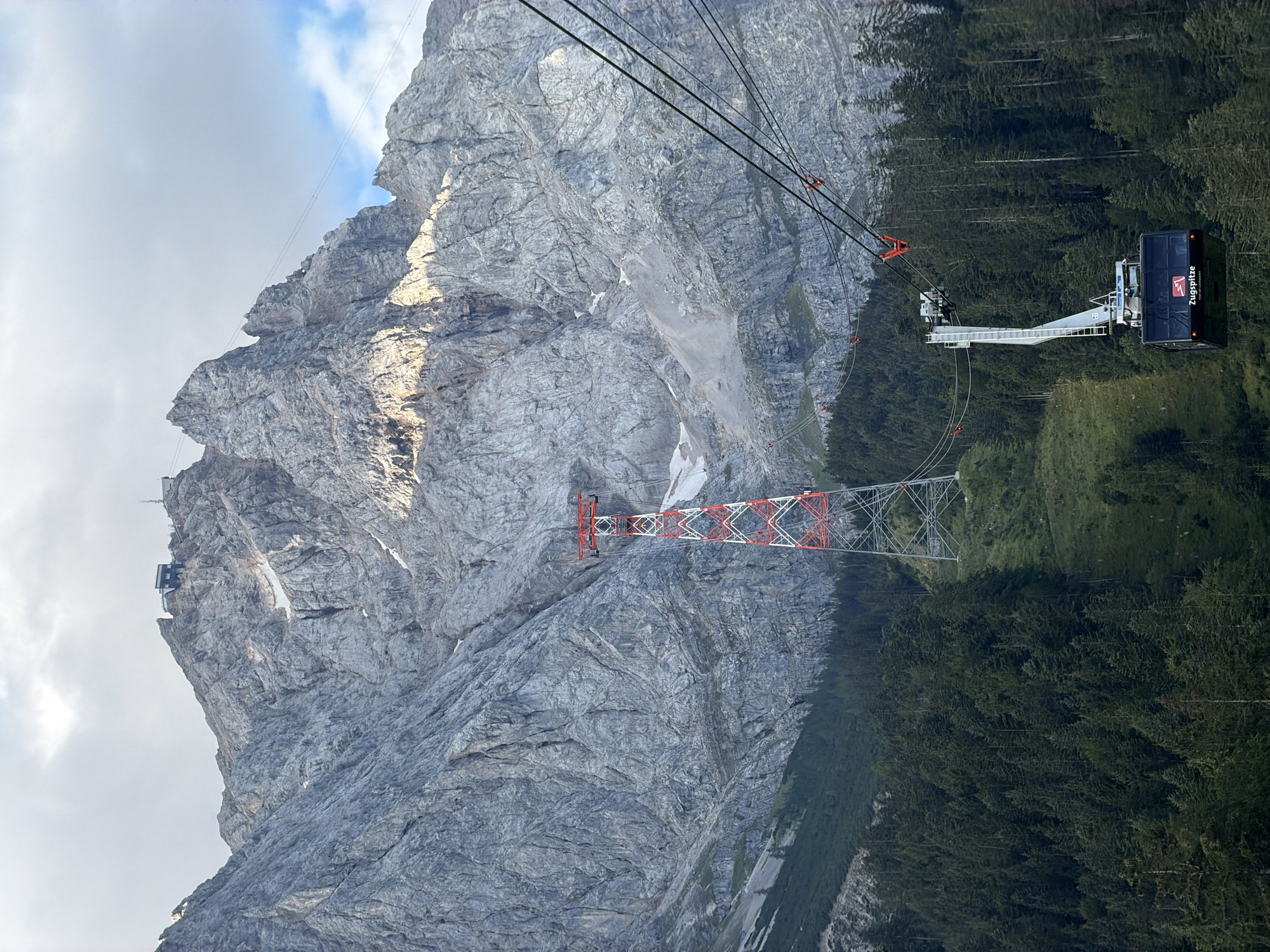 This image captures a dramatic mountain landscape in Grainau, Germany, likely the Zugspitze region, dominated by a towering grey, rocky mountain. A striking red and white pylon stands tall on the mountainside, supporting cables of a cable car system. In the foreground, a dark cable car cabin with the label "Zugspitze TOP OF GERMANY" is suspended mid-air, ascending towards the summit. The mountain face is rugged and textured, with patches of snow visible in crevices. A dense, dark green forest covers the lower slopes of the mountain, creating a stark contrast with the barren rock above. The sky is overcast with scattered clouds, hinting at either the early morning or late afternoon light. No people are visible in the image, and the scene is serene and majestic.