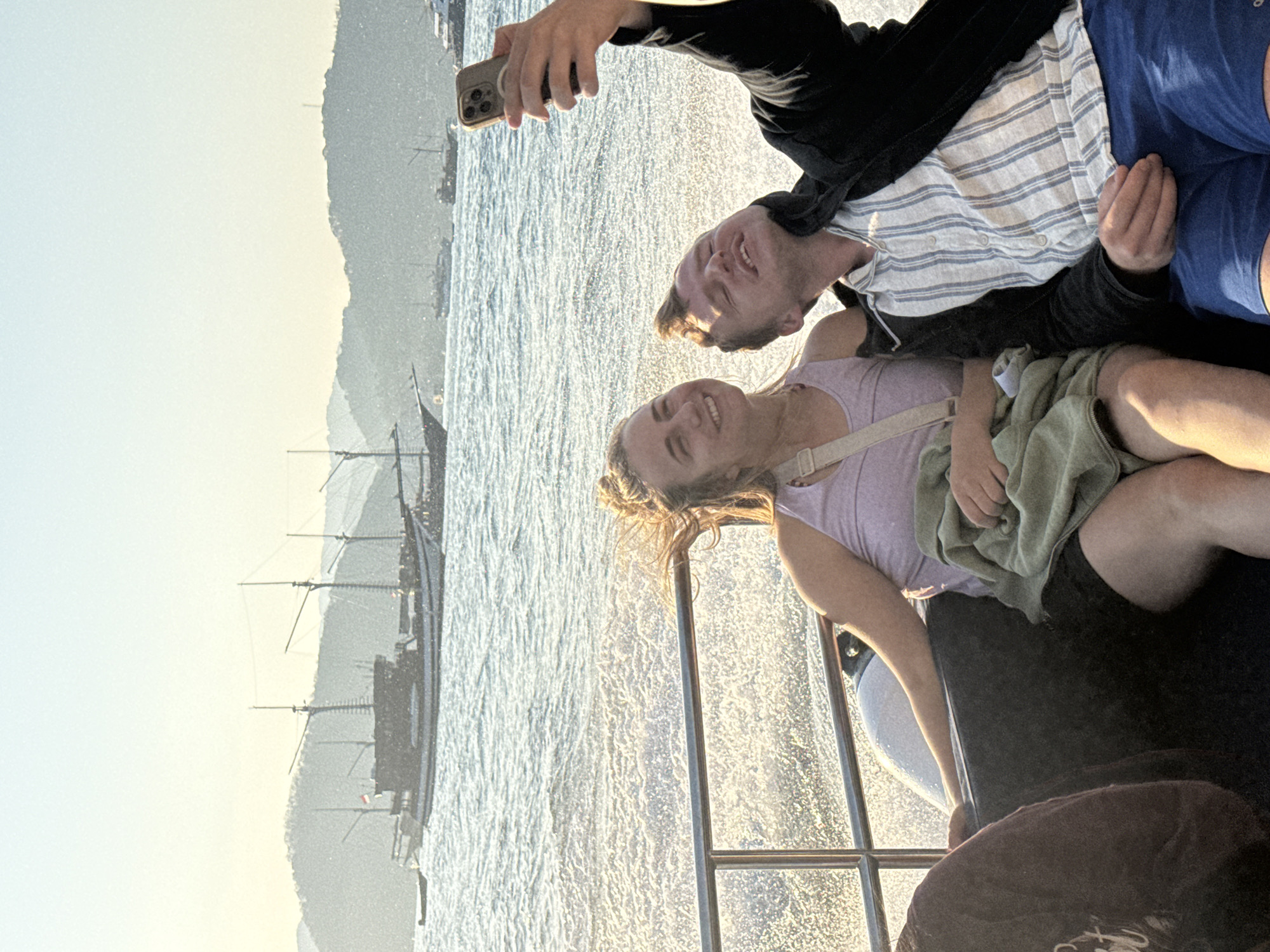 The image shows a young couple taking a selfie on a boat in Labuan Bajo, Indonesia. The man, on the right, is holding up a smartphone to take the picture. He is wearing a black jacket over a striped shirt and blue shorts. The woman, on the left, is wearing a pink tank top and black shorts. She is smiling and leaning towards the man. They are sitting on a boat with a metal railing in front of them. The water behind them is choppy and reflects the sunlight. In the background, there are several other boats, including a large traditional sailing ship, and a hazy silhouette of mountains. The sun appears to be setting, casting a warm glow over the scene.