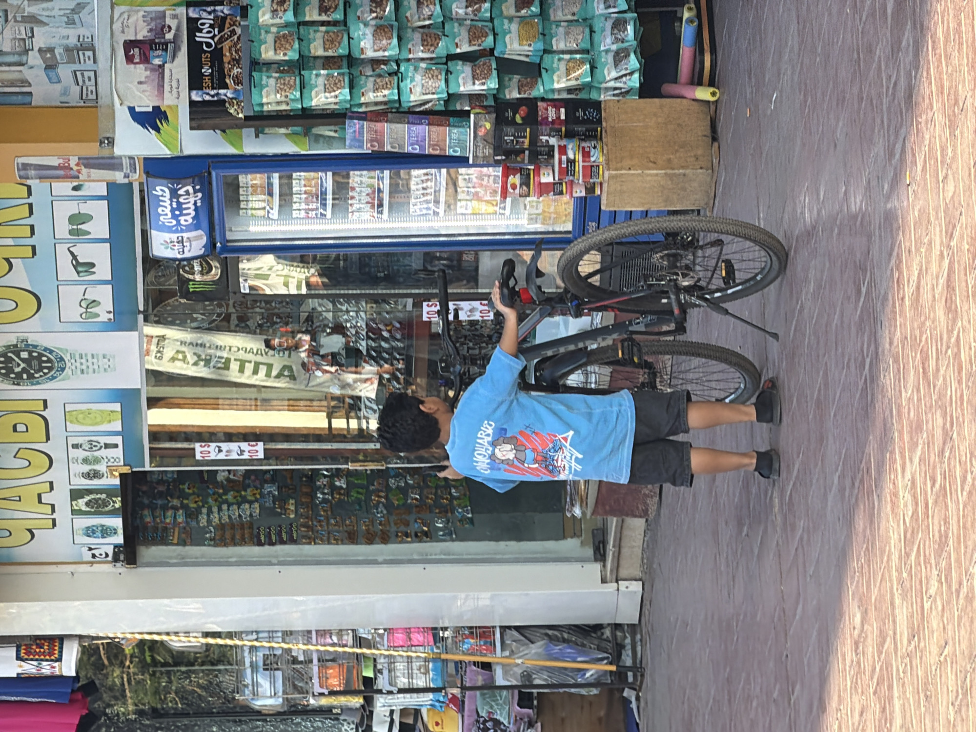 This media file shows a young boy standing next to a bicycle in an outdoor setting. He is wearing a light blue t-shirt with a graphic design on the back and dark shorts. The bicycle is a dark color with a visible wheel and handlebars.
The scene appears to be in a marketplace or shop area. Behind the boy and the bicycle, there is a storefront with various items displayed, including packaged food products, drinks in a refrigerator, and what look like watches or accessories. Some signage is visible, though not all of it is legible. One sign has yellow lettering with black outlines that reads "СПЕЦИАЛЬНЫЕ". Another sign, partially visible, has Arabic script. There are also price tags that say "10 $" and "10 €".
The ground is paved with red bricks. The overall lighting suggests it might be daytime, possibly late afternoon given the angle of the shadows. The boy's posture is relaxed as he stands with his phone in hand. The environment seems to be a busy commercial area, possibly a tourist destination given the presence of multiple languages on signs. The context provided indicates the location is Sharm El Sheikh, Egypt.