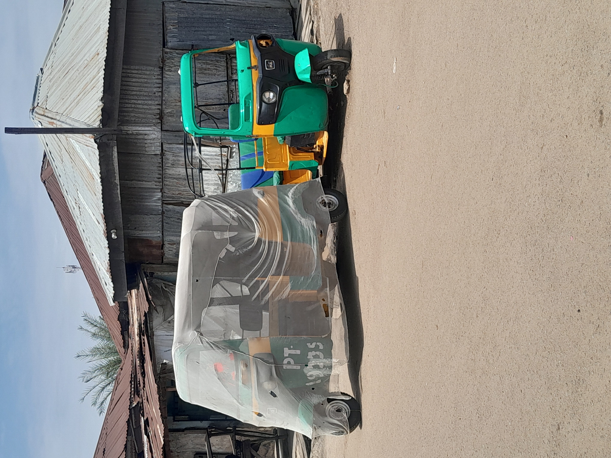 The image shows two auto-rickshaws (tuk-tuks) parked on a paved surface alongside a building in Jos, Nigeria. One tuk-tuk is covered in a translucent plastic sheet. The other is green and yellow and is not covered. The building is constructed with corrugated metal sheets. There is no evidence of human activity in the frame. The weather appears sunny, and the light is casting distinct shadows.