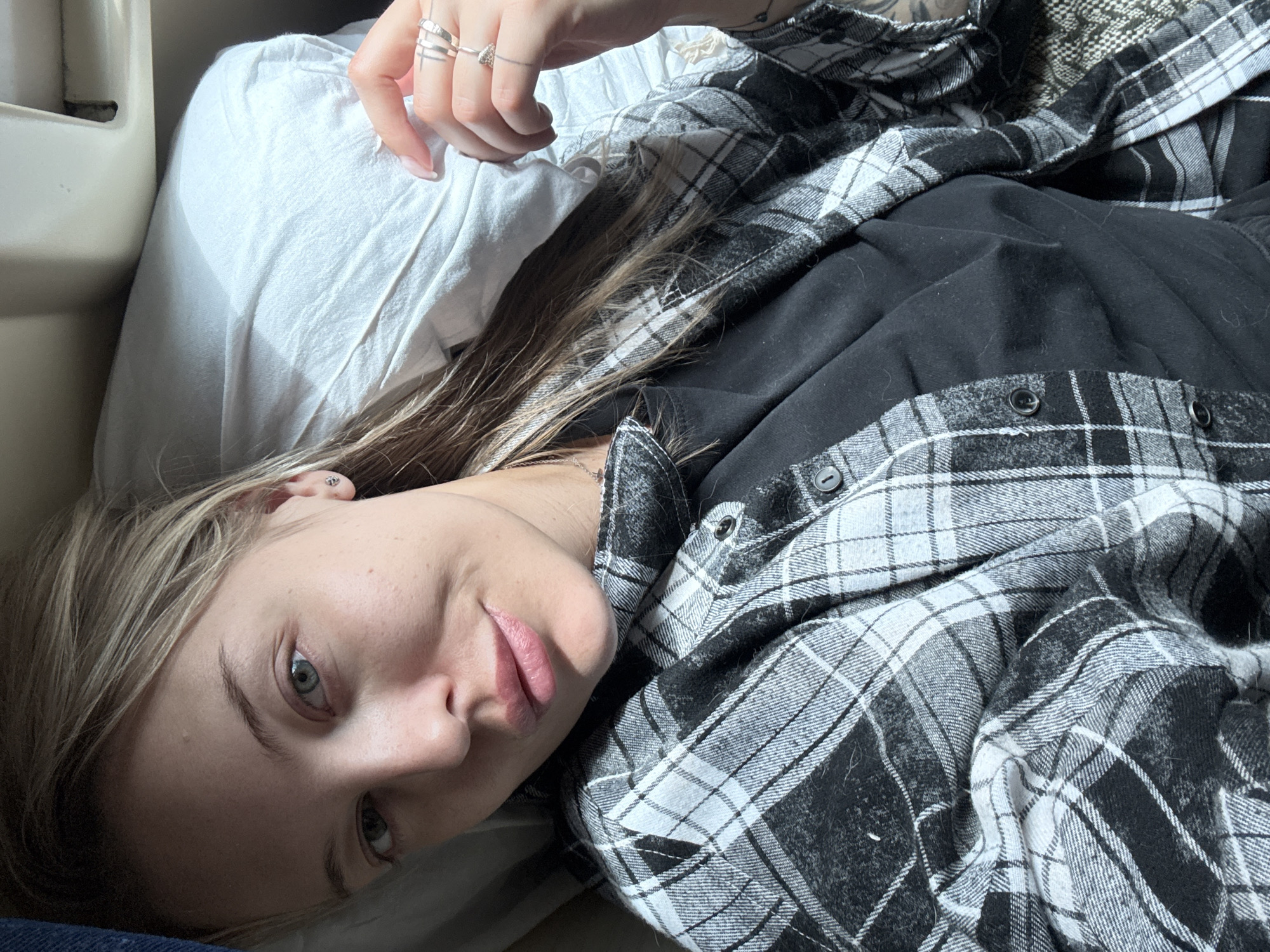 A young woman lies on a white pillow, wearing a black t-shirt and a black and white plaid flannel shirt. She looks towards the camera with a slight smile. She has blonde hair and light eyes. Her hand, adorned with multiple rings, rests near the pillow. No details suggest the location is Bada, Russia.