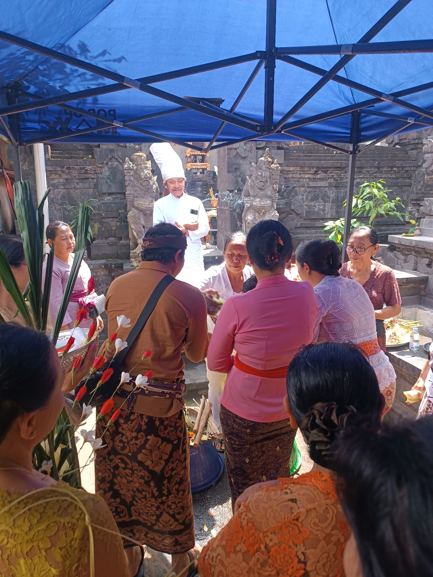 The photograph depicts a ceremony taking place outdoors, likely at a temple or traditional Balinese compound in the city of Tabanan, Indonesia. A group of individuals, predominantly women, are gathered. In the center, a man wearing a tall white chef's hat and a white uniform stands, appearing to be leading or participating in the ceremony. He has a mustache and is looking downwards. To his left, a woman in a light purple outfit with a sash is visible. Several other women in traditional Balinese attire, including sarongs and blouses, are positioned around the central figures. One woman in the foreground is wearing a brown long-sleeved shirt and a patterned sarong. Another woman in the foreground is wearing a brightly colored orange lace top. The scene is partially covered by a blue canopy supported by dark poles. In the background, there are stone structures with intricate carvings, indicative of Balinese architecture, and lush green plants. A basket containing what appears to be offerings or floral arrangements, including red and white flowers, is also visible. The ground is paved with a mix of stone and dirt. The overall atmosphere suggests a cultural or religious event.