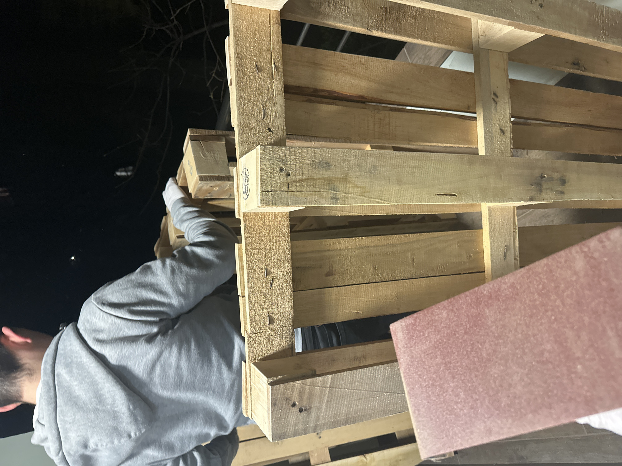 A man in a grey hoodie is stacking wooden pallets, seemingly outdoors at night. The area is dimly lit with a dark sky visible. A pinkish-red rectangular object is visible near the pallets. The location cannot be determined to be Sofia, Bulgaria based on the image.