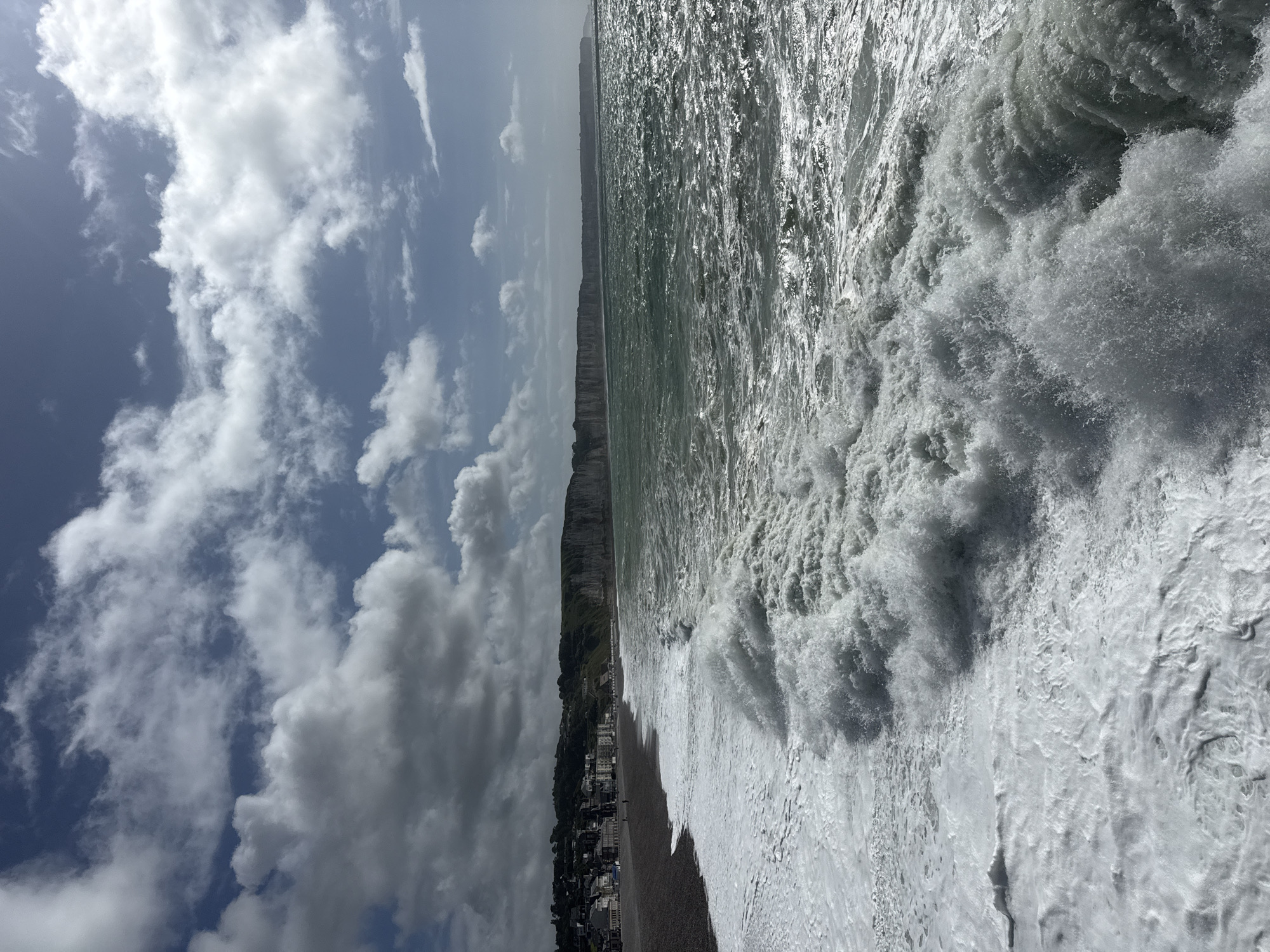The image presents a dramatic coastal scene at Fécamp, France, captured under dynamic weather conditions. The photograph is oriented vertically, but depicts a landscape view.
The sky, occupying the upper left portion (if correctly oriented), is a mix of bright blue patches and large, fluffy cumulus clouds, some appearing white and others grey, indicating a partly cloudy but bright day.
In the mid-ground, a striking feature is the long stretch of light-colored cliffs that extend horizontally into the distance along the coastline. Below these cliffs, a coastal town is visible, with numerous buildings nestled along the shore, characteristic of a seaside settlement.
The dominant feature of the scene is the powerful, turbulent sea, which fills the right side and foreground of the image. Large, foamy waves crash vigorously towards the shore, their white crests contrasting with the darker, shimmering surface of the water, which reflects the bright sunlight intensely. The high reflection suggests the sun is high in the sky, likely mid-day. The intense wave action indicates very rough sea conditions. A pebble beach is visible where the waves break and recede.
The overall impression is one of natural power and grandeur. There are no discernible people or specific objects beyond the town's buildings, and no visible text.
