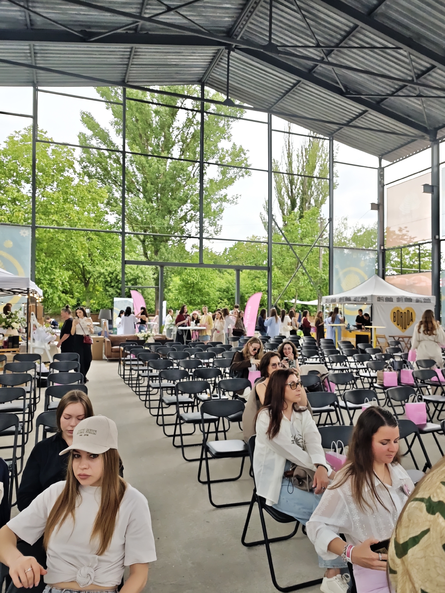 This image depicts a daytime event taking place within a large, open-sided structure with a corrugated metal roof supported by exposed black metal beams and trusses. The background features a panoramic view of lush green trees and a bright, overcast sky. The event is located in Vinnytsia, Ukraine, identified by a white canopy tent in the background displaying a yellow heart logo with the word "BORODA."
In the foreground, numerous black folding chairs are arranged in rows on a concrete floor. On the left, a woman with light brown hair wears a white baseball cap and a white t-shirt tied at the waist, looking towards the viewer. A silver ring is visible on her left ring finger. Behind her, another woman with light brown hair wears a black long-sleeved top and looks downwards. On the right, a woman with dark hair wears a white textured long-sleeved shirt and looks down at a handheld electronic device. A pink paper bag is visible next to her. Further right, a woman with dark hair and glasses, dressed in a white collared shirt and light blue jeans, is seated, looking towards the left.
The mid-ground contains additional rows of black folding chairs, many occupied by women. Several pink paper bags are placed on or adjacent to the chairs throughout the seated area.
The background displays active event stalls and groups of individuals. On the left, a white canopy tent serves as a vendor booth with various items displayed and a woman in a black shirt behind a counter. Towards the right, the aforementioned white canopy tent with the "BORODA" logo shows two men behind a counter, appearing to serve customers. A woman in a light-colored long coat is walking away from this tent. In the deeper background, a brown leather sofa, additional tables, and smaller groups of people are visible, indicating areas for socializing or product displays. Large fabric banners with abstract designs, including a blue banner with a white star-like emblem on the left, are affixed to the structural supports. The event appears to be a gathering or market with both a formal seating area and informal vendor zones.