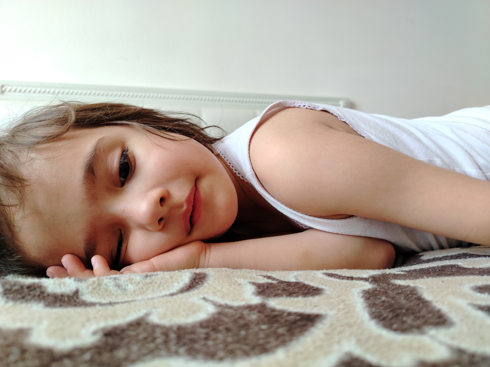 A young child with brown hair and fair skin lies on a patterned bedspread. The child is wearing a white tank top. One eye is open, and the child is slightly smiling, while resting their cheek on their hand. The background is a plain, light-colored wall. There is no evidence to suggest the location is the city of Noviy, Russia.