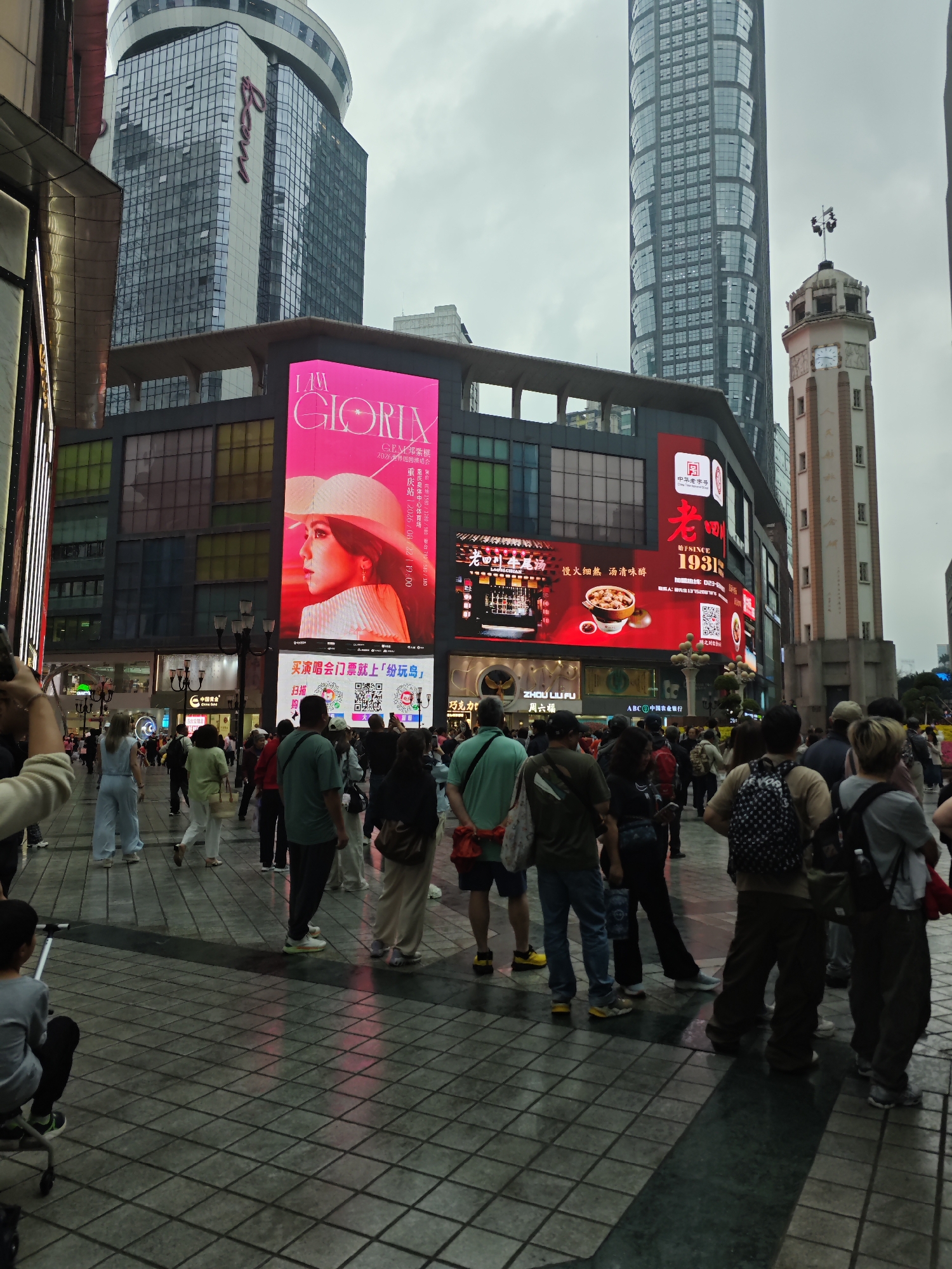 A crowded pedestrian area in Yuzhong District, China, features several prominent commercial buildings. A large screen displays an advertisement for a GEM concert and another screen shows an advertisement for a food establishment that has been around since 1931. People are standing and walking. A clock tower is visible in the background.