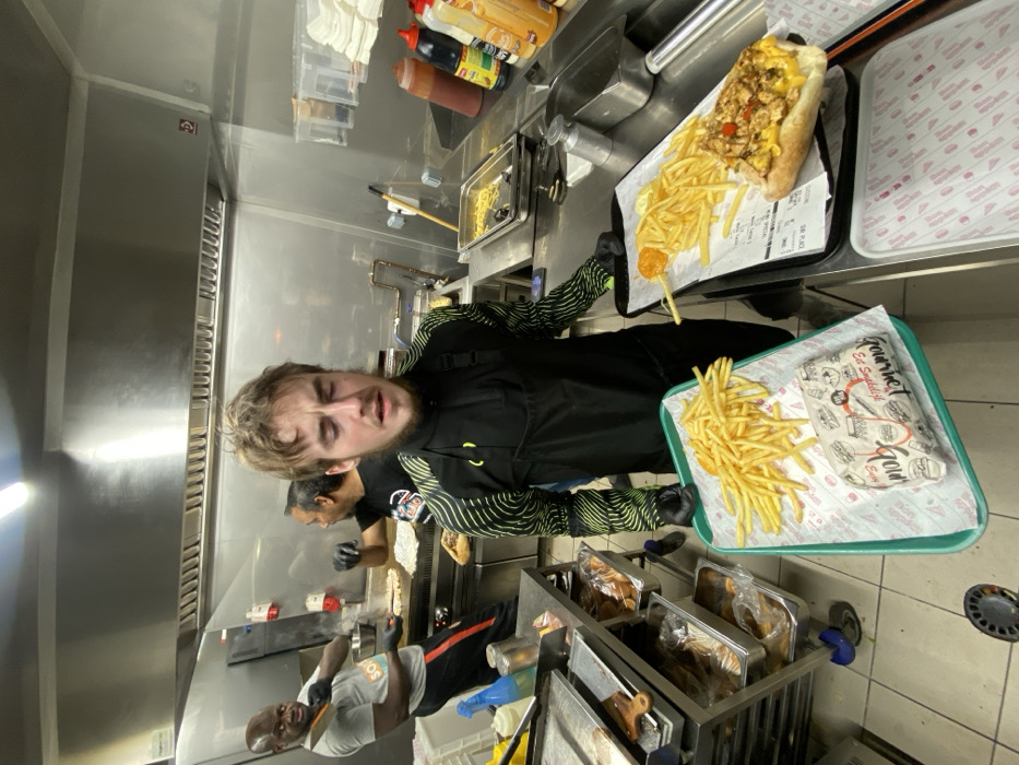 Inside a commercial kitchen, a fair-skinned person holds trays of fries and sandwiches. Two other workers are present. One is slicing food. The kitchen features stainless steel surfaces, fryers, and condiment bottles. Trays are lined with paper, displaying fries and sandwiches. The scene is brightly lit with fluorescent lighting.