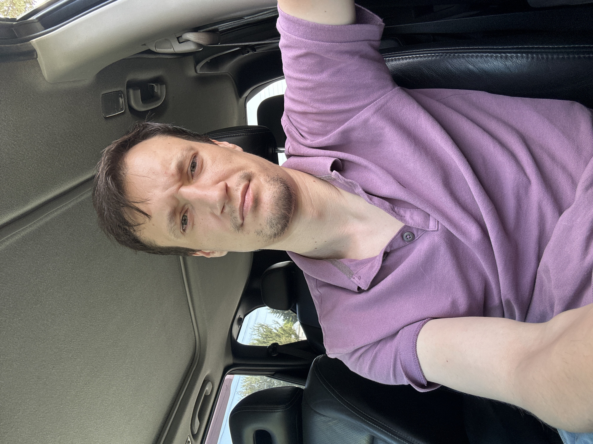 A man with medium-length brown hair and light skin is seated in the driver's seat of a car. He is wearing a purple polo shirt and looking at the camera with a slight frown. The interior of the car is visible, including black leather seats, the car's headliner, and a sunroof. Through the car windows, trees and a building can be seen, suggesting an outdoor location, possibly in a suburban or rural setting.