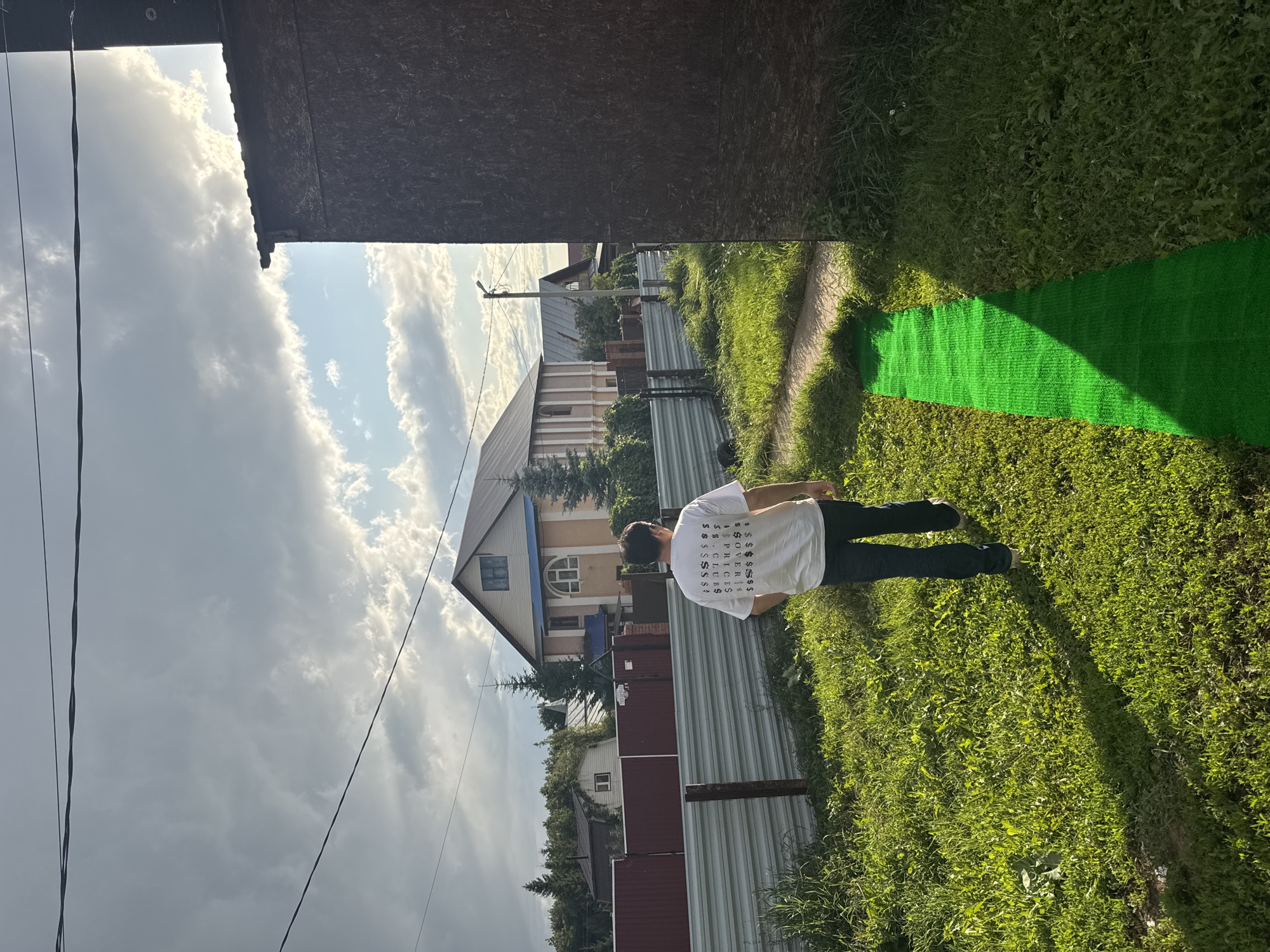In Nikolaevka, Russia, a person is walking away from the camera on a grassy area. They are wearing a white t-shirt with black text and dark pants. The t-shirt has a graphic design consisting of multiple dollar signs and the words "PRICES", "CLUBS", and "SOVER". In the background, there are houses, a fence, and trees. The sky is cloudy with some patches of sunlight. A bright green mat or rug is laid out on the grass to the right of the person.