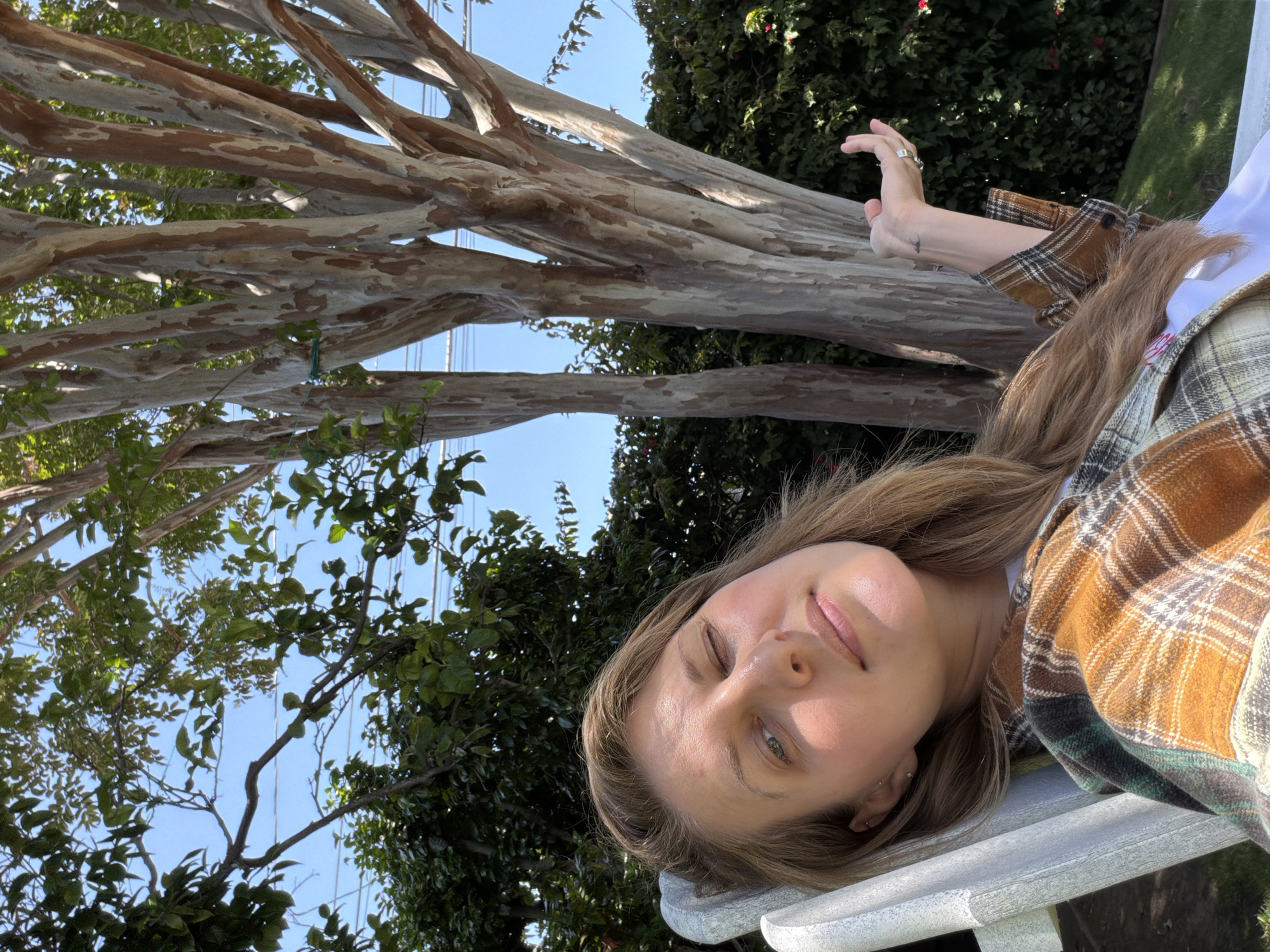 This image depicts a female individual outdoors in Santa Monica, United States of America, under bright natural light. The subject, a Caucasian female with long, light brown hair, is reclining in a light-colored outdoor chair. Her left eye is closed, indicating a wink or squint, while her right eye is partially open and directed towards the viewer. She is wearing a brown, green, and white plaid flannel shirt over a white undergarment. A small dark tattoo, resembling a wave or horizon line, is visible on her inner left wrist. She wears multiple rings on the fingers of her raised left hand. Two small stud earrings are visible in her left ear.
The background features the distinct, multi-trunked, exfoliating bark of a crape myrtle tree (Lagerstroemia), displaying patches of gray, white, and reddish-brown. Surrounding the crape myrtle are various dense green trees and shrubs, some with visible red blooms. Above the foliage, a clear blue sky is visible, with several horizontal utility lines traversing the upper portion of the frame. The immediate ground level visible appears to be green grass.