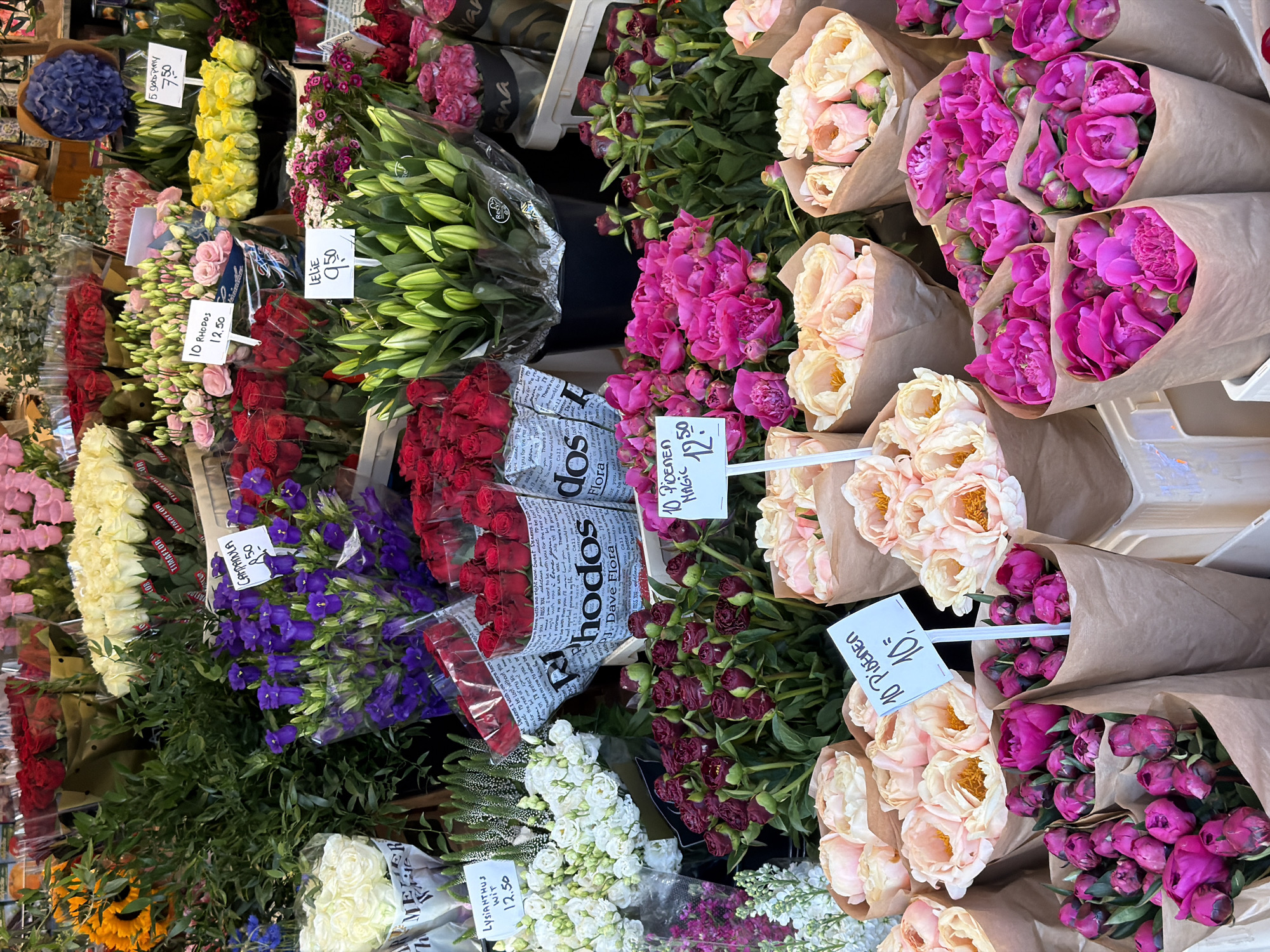 The image depicts a flower stall in Amsterdam, Netherlands. The stall showcases a variety of flowers, including peonies (both pink and cream), roses (red and pink), lilies, campanula (purple), lysianthus (white), and sunflowers. The flowers are arranged in bouquets and bundles, with price tags indicating the cost. The price tags are in Dutch. The flowers are displayed in a row, creating a colorful and vibrant scene. Some of the flowers are wrapped in paper or plastic, while others are displayed in buckets. The stall is likely located in a busy market or street.