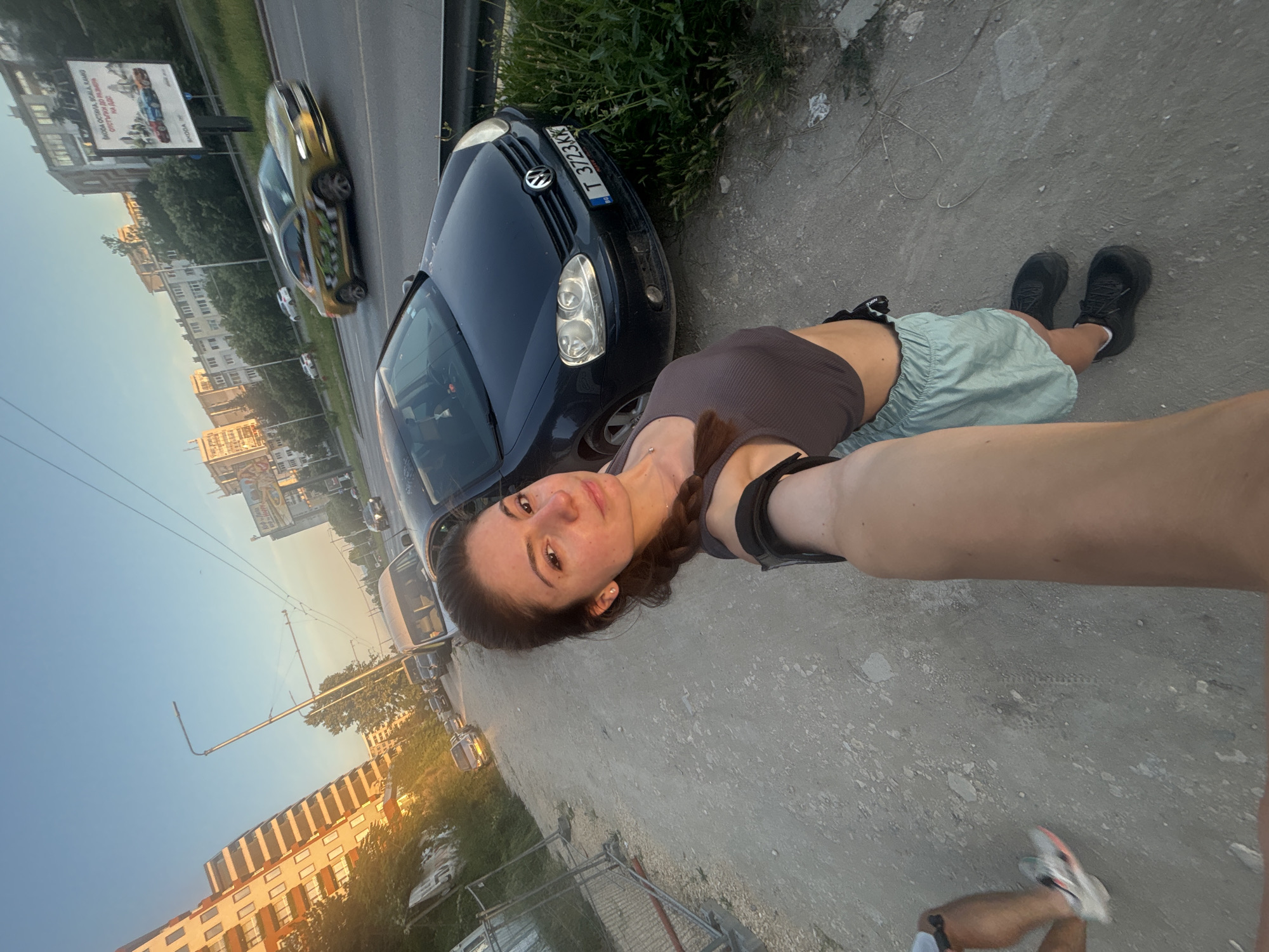 A young woman is taking a selfie on a dirt path next to a busy road in the city of Varna, Bulgaria. She is wearing a grey crop top and light blue shorts with black sneakers. Her hair is dark brown and pulled back into a braid. In the background, there are several cars driving on the road, apartment buildings, and a billboard. The sun is setting, casting a warm glow over the scene. The woman's left arm is extended, holding the camera. To the left of the woman, a person's legs are visible, with one leg in motion as if running or walking, wearing a white and orange running shoe. The environment appears to be urban with signs of ongoing construction on the left side of the image, indicated by metal fencing.