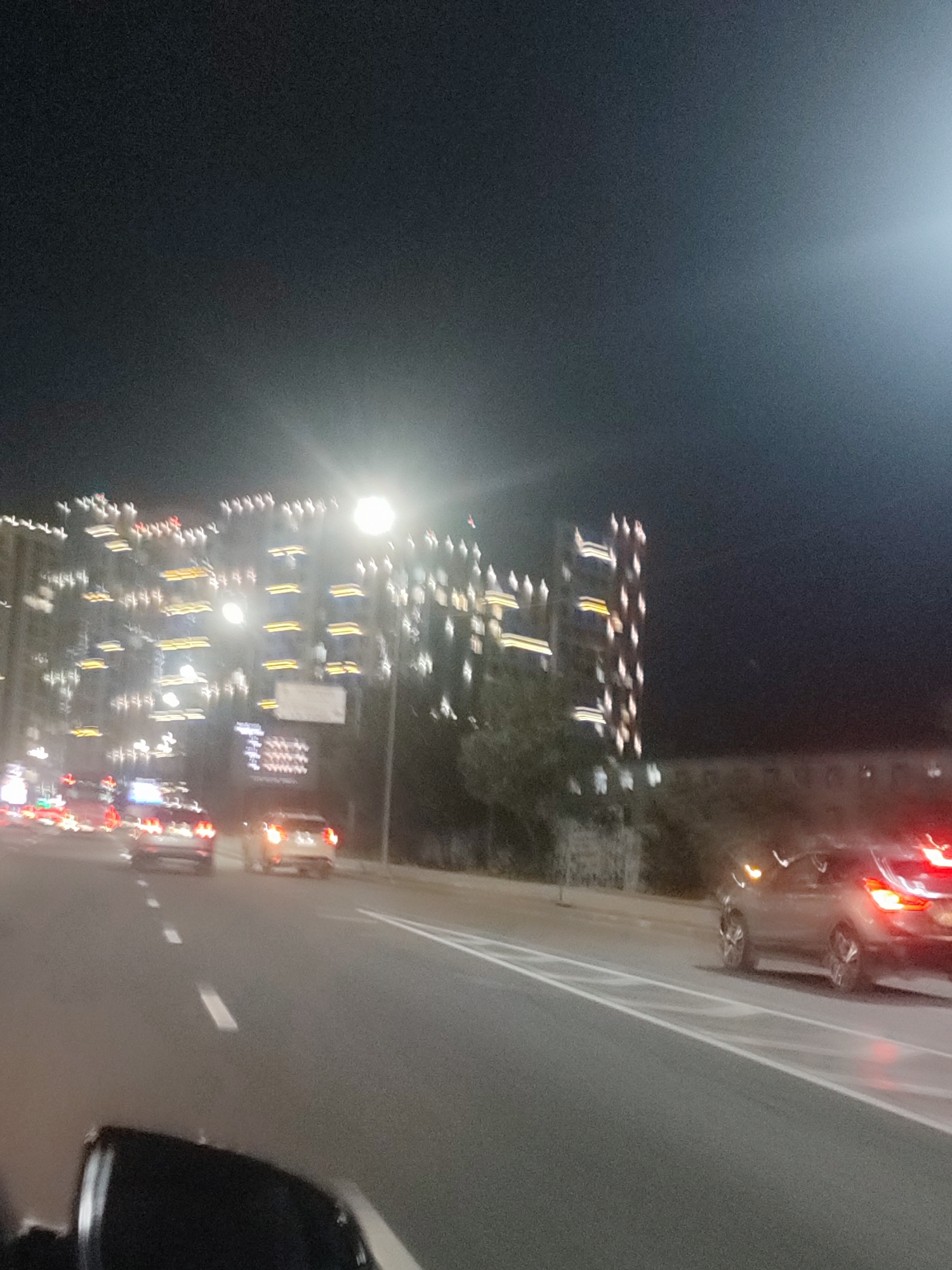 The image depicts a nighttime street scene in Almaty, Kazakhstan. A multi-story building illuminated with numerous lights is visible on the left side of the frame. Cars are driving on a road in the foreground, with their headlights and taillights creating streaks of light due to the long exposure. One car, a dark sedan, is visible in the right lane, and its brake lights are illuminated. The sky is dark, suggesting it is nighttime. The scene appears to be on a relatively wide road, possibly a highway or major thoroughfare within the city. The image is slightly blurred, likely due to motion or low light conditions.