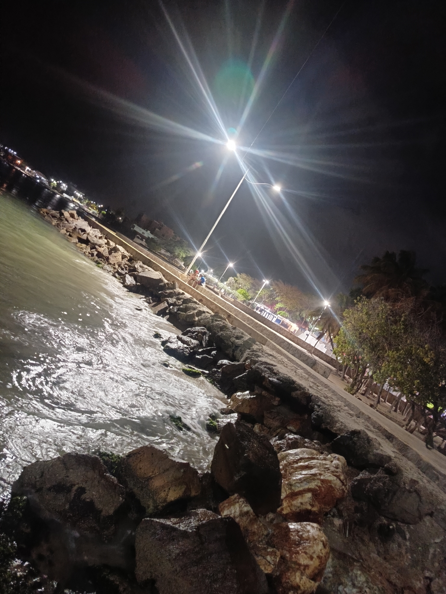 Captured at night in Juan Griego, Venezuela, the image showcases a waterfront scene. The foreground features large, dark rocks, partially submerged in water. The water reflects light, giving it a shimmering appearance. Adjacent to the rocks is a concrete walkway, illuminated by streetlights. Along the walkway, there are sparse trees. People are visible in the distance, near the streetlights. The background is dark, with a few lights from buildings across the water. The overall composition has a bright streetlight shining directly towards the camera, creating a starburst effect.