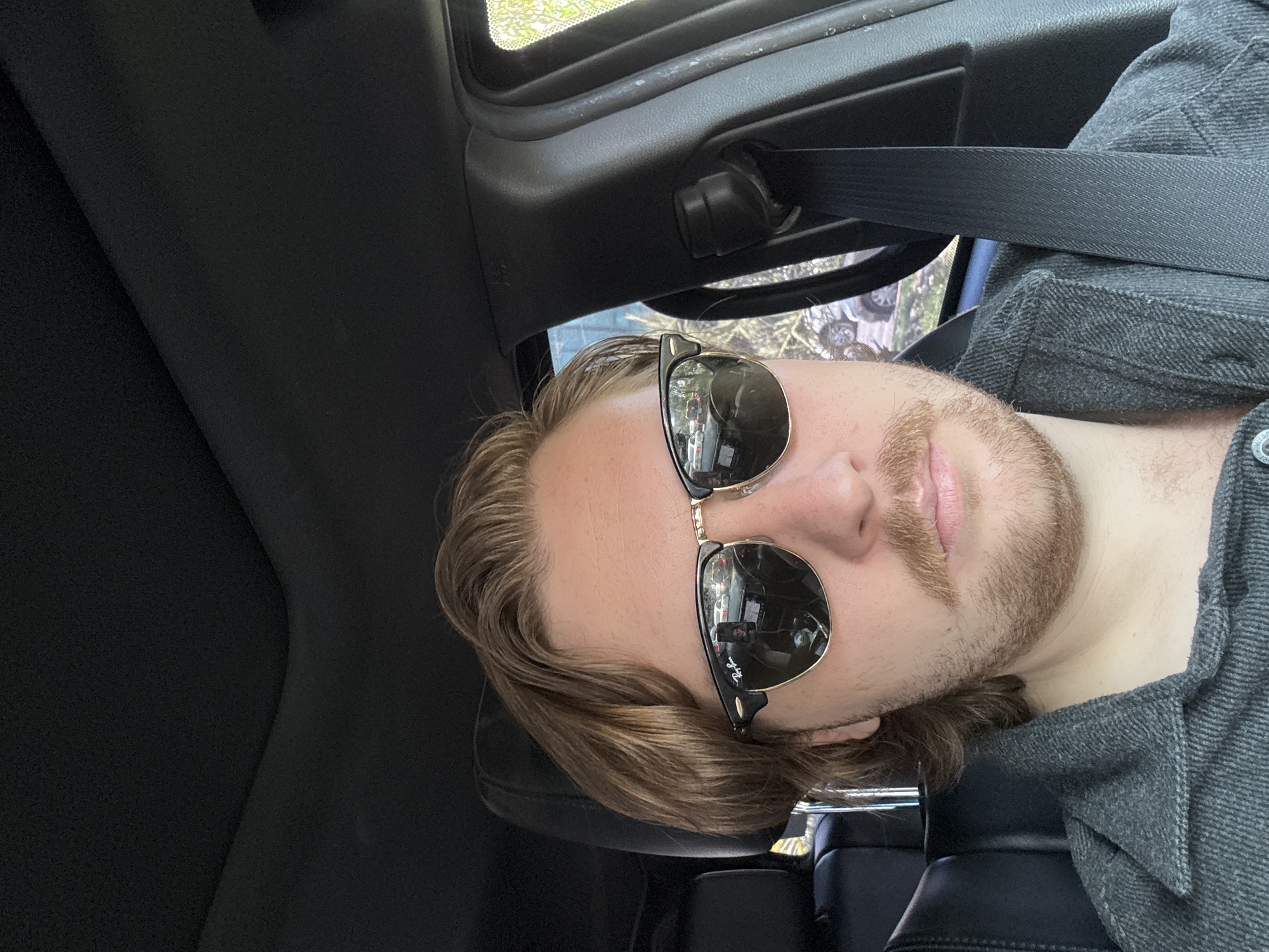 A close-up, rotated image shows a person (male) with light brown hair, a mustache, and a beard, wearing black and gold Ray-Ban Clubmaster style sunglasses. The person is situated inside a vehicle, likely in the driver's or front passenger's seat, indicated by the visible black car interior, headrest, door frame, and seatbelt across their chest. They are wearing a gray collared shirt or jacket.
Reflections are prominent in the sunglasses. The left lens (from the viewer's perspective) clearly shows the front of a white or light-colored vehicle, possibly a truck or SUV, with its lights visible. The right lens reflection appears to show a person holding a device, suggesting the image is a selfie or taken by someone very close to the subject.
Through the car window, blurred green and brown vegetation, indicative of trees or foliage, can be seen, implying an outdoor setting during the daytime. The overall lighting suggests it's a bright day. The text "Ray-Ban" is visible on the sunglasses. The location is Mexico City, Mexico.