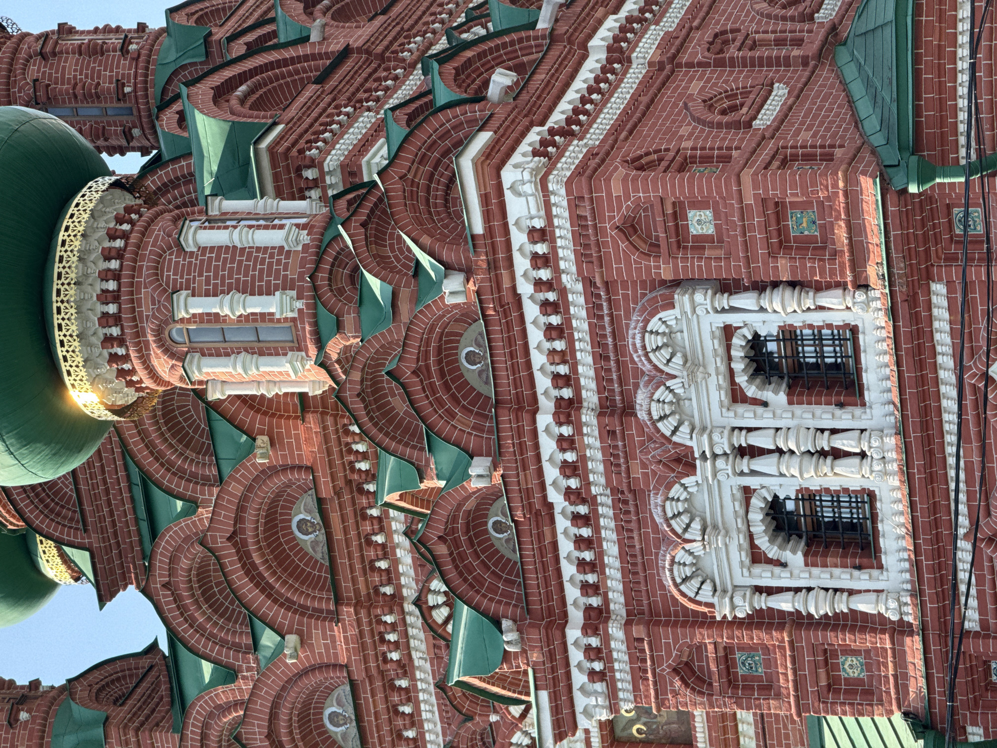 The image shows a close-up view of St. Basil's Cathedral in Moscow, Russia. The building is constructed of red brick with white architectural detailing, green roofs, and ornate patterns. A portion of a green dome is visible, reflecting sunlight. The perspective is from below, looking upwards at an angle. Architectural elements such as decorative arches, windows with bars, and rows of white detailing are prominent. The sky is visible as a small blue patch.