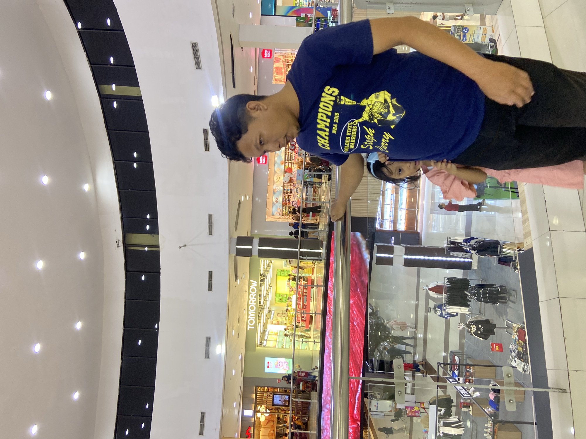 In an indoor shopping mall, a man in a dark blue t-shirt with a "Champions NBA 2015 Golden State Warriors Steph Curry" graphic stands at a railing. He is looking to his left. Partially obscured behind him, a young girl in a pink dress peeks out, also looking towards his left. The man's left hand rests on the railing. The background shows multiple levels of the mall with storefronts, including one labeled "TOMORROW." The environment is well-lit with overhead lights and displays. The scene is consistent with a shopping mall in Sidoarjo, Indonesia.