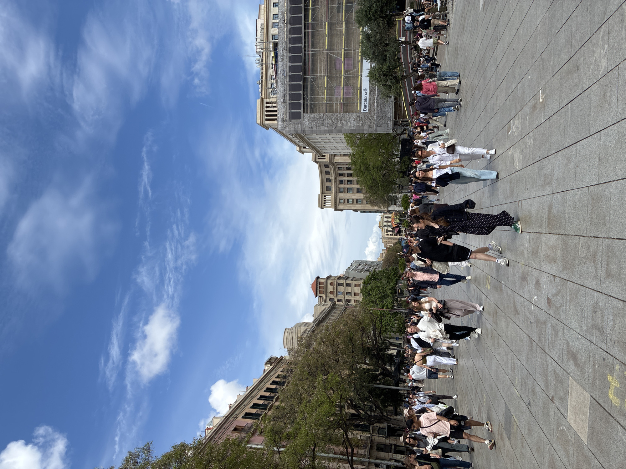 The image shows a public square in Barcelona, Spain, likely Plaça de Catalunya, densely populated with people. The square is paved with large gray stones. Buildings with varied architectural styles surround the square, with one under construction and covered in scaffolding. The sky is blue with scattered white clouds. People are walking, standing, and interacting with each other, suggesting a vibrant urban scene. The overall atmosphere appears busy and lively.