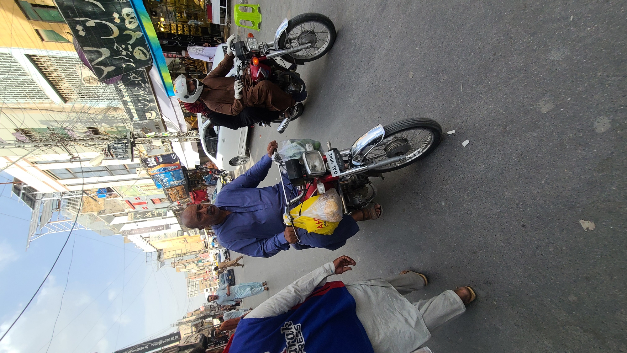 Here is a detailed description of the image:
**Scene:**
The image appears to be taken on a busy street in Quetta City Tehsil, Pakistan. The street is lined with buildings and various commercial establishments with signage in Urdu and English.
**People:**
* A man is sitting on a red motorcycle, holding a yellow plastic bag with a loaf inside. He wears a blue tunic.
* Another man is riding a motorcycle wearing a brown outfit and a white helmet.
* A man is lying on the street wearing traditional shalwar kameez.
* Several other people are seen walking on the street in the background, mostly wearing traditional Pakistani clothing.
**Objects:**
* Two motorcycles are prominent, one red and one brown.
* A yellow plastic bag with a loaf inside.
* Various storefront signs with text including "JEWLERS" and "PRESS".
* A white car is parked on the side of the road.
**Environment:**
* The image is taken in daylight.
* The street is paved and shows signs of wear and tear.
* The buildings have a mix of old and new architectural styles.
**Location Clues:**
The presence of signage in Urdu and the clothing styles suggest a South Asian location. The license plate on the red motorcycle says "QAL 9042" which corresponds to vehicles in the Quetta district.