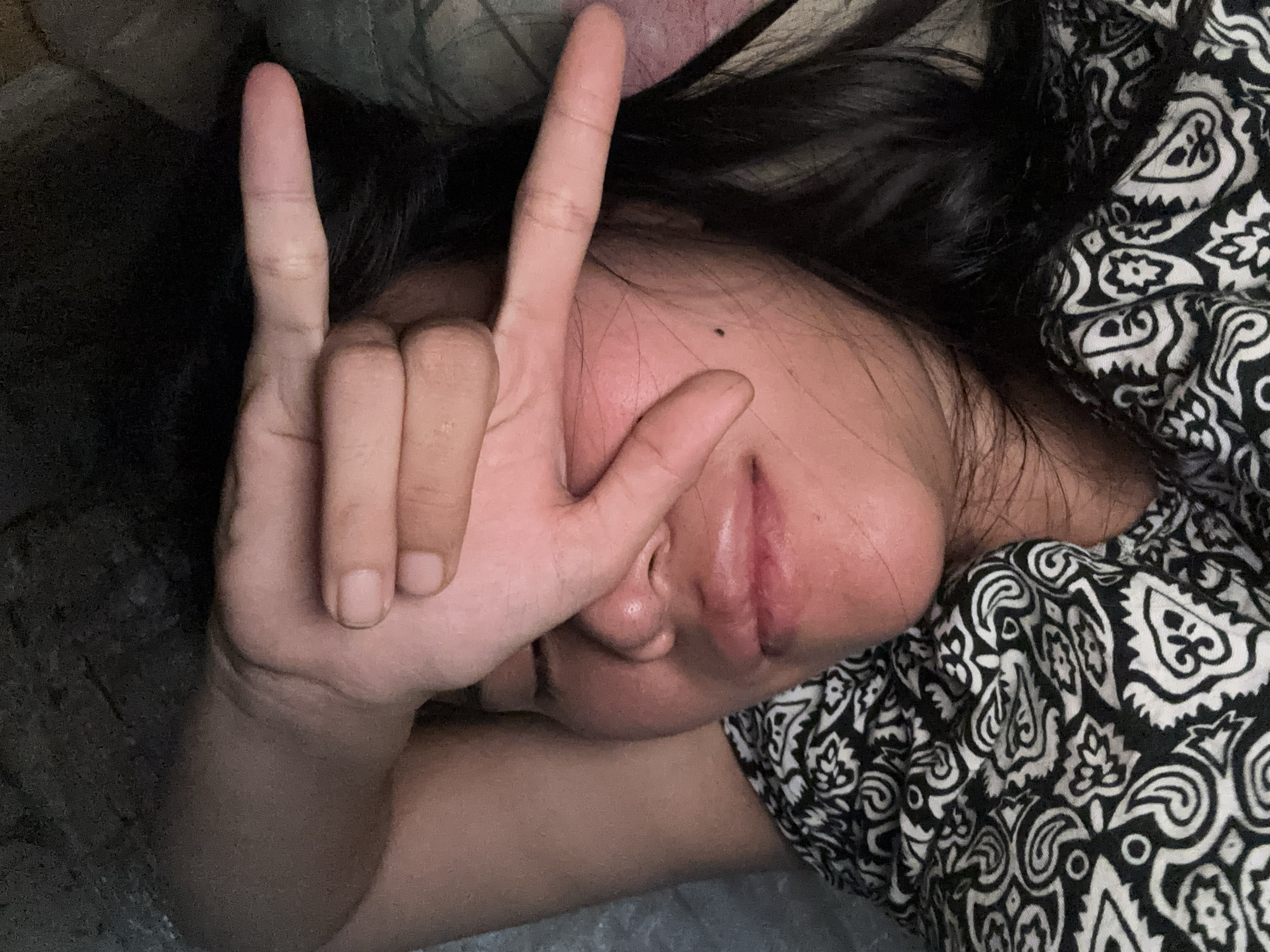 A person lies on their back with their eyes closed, their face partially obscured by their left hand, which is positioned over their forehead and eyes. The hand forms a gesture with the index and middle fingers extended, and the thumb, ring finger, and pinky finger curled inward. The person is wearing a black and white patterned shirt. The background appears to be a dimly lit interior, possibly a bedroom, with textures suggesting a textured wall or fabric. The person's skin tone is fair, and they have dark hair. The lighting is warm and appears to be coming from a single source, casting shadows on the face and creating a soft glow. The image is a close-up, focusing on the person's face and upper body. There is no indication of the location being in East Jakarta, Indonesia, or any specific city.