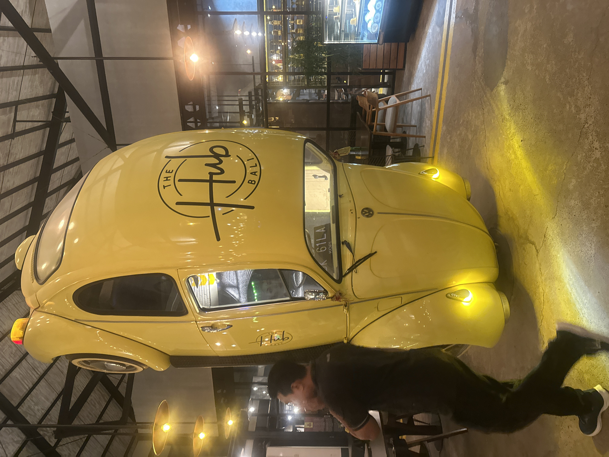 The image shows a yellow Volkswagen Beetle car suspended upside down indoors. The car is decorated with a logo that says "The Hub Bali." A man is walking past the car on the left side of the image. The scene appears to be inside a cafe or restaurant in Denpasar, Indonesia. There are tables and chairs visible in the background, as well as a display case with pastries. The lighting is warm and atmospheric, with yellow lights casting shadows on the floor.