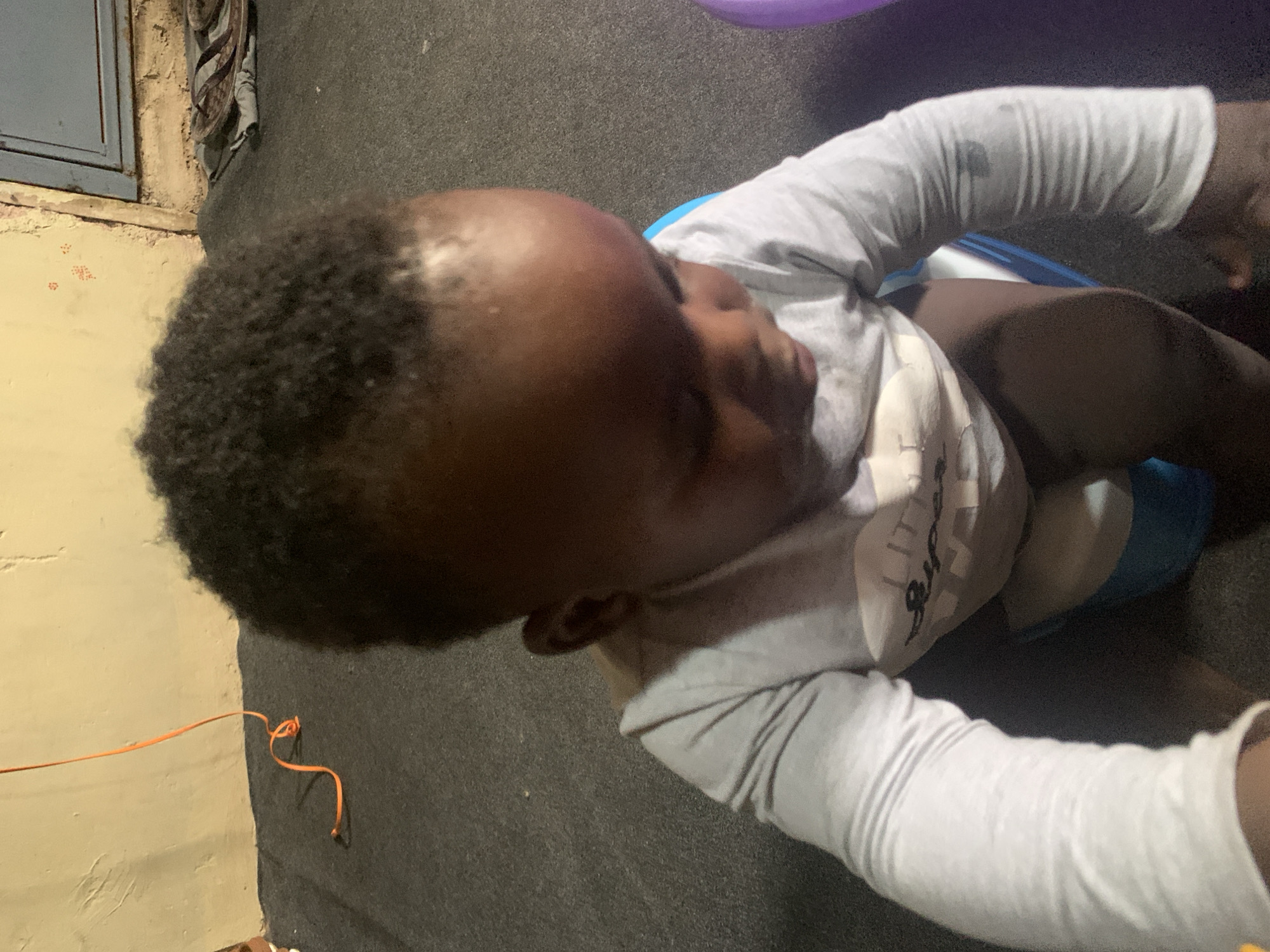 A young child, likely a toddler, is seated on a blue and white potty training seat indoors. The child is wearing a light gray long-sleeved shirt with a graphic on the front. The child's head is tilted down, and their facial features are obscured. The background is indistinct but appears to be a room with a textured floor and a wall. An orange cord is visible on the left side of the image, and a pair of flip-flops are on the floor in the upper right portion of the frame. The lighting is dim.