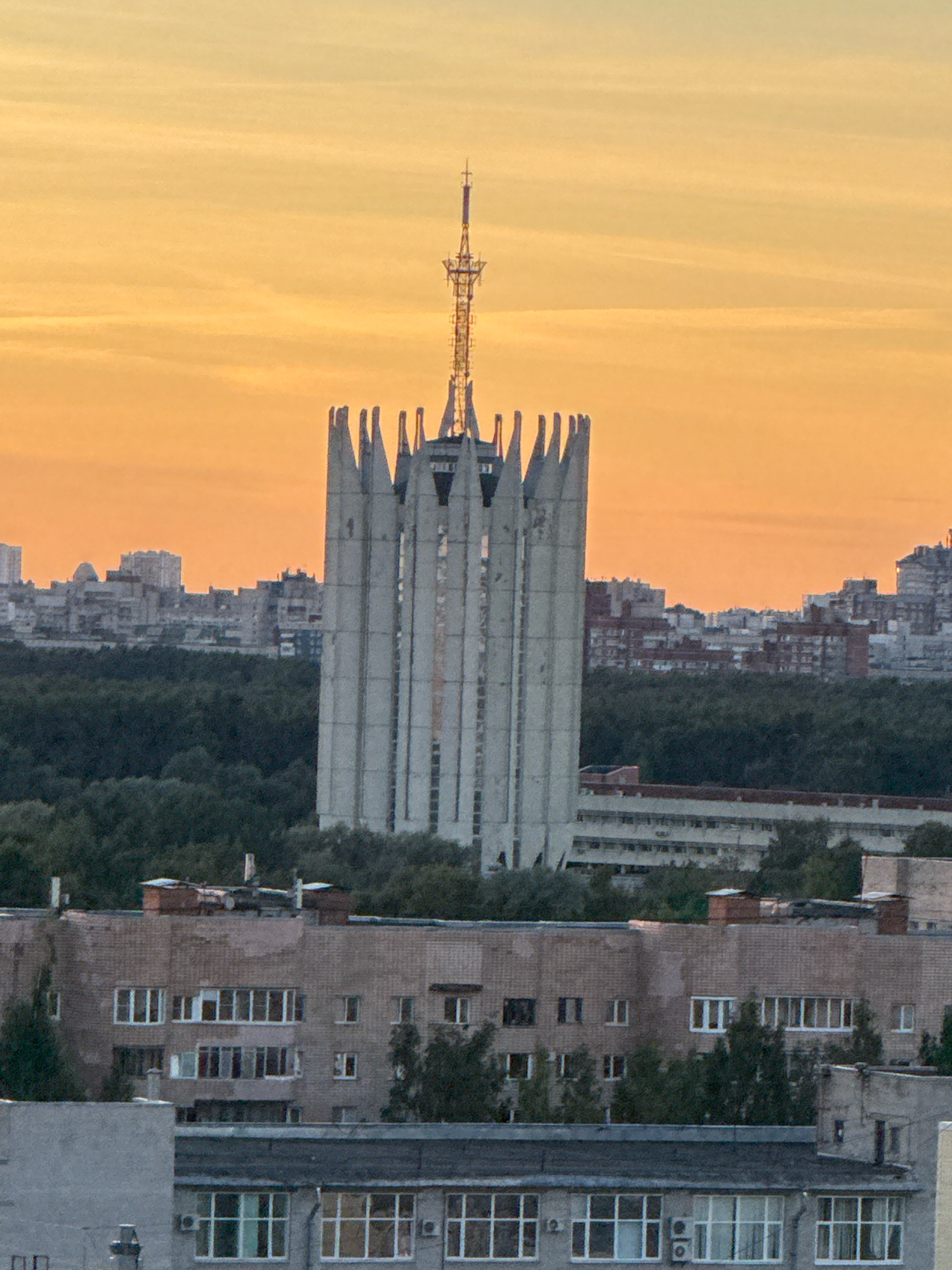 The image is a medium-angle, slightly elevated shot of a city skyline at sunset. The sky is a gradient of orange and yellow hues. In the foreground, there are several apartment buildings constructed from brick and concrete, exhibiting typical Soviet-era architecture with numerous windows. Behind these buildings, a large, Brutalist-style concrete tower with a distinctive crenellated top stands prominently. This tower is topped with a communication antenna. Beyond the tower, a dense line of dark green trees forms a natural barrier, separating the foreground from a more distant cityscape of various buildings and structures. The overall impression is one of an urban environment at dusk, with a mix of residential and utilitarian architecture. The location is Sinyavino, Russia.
