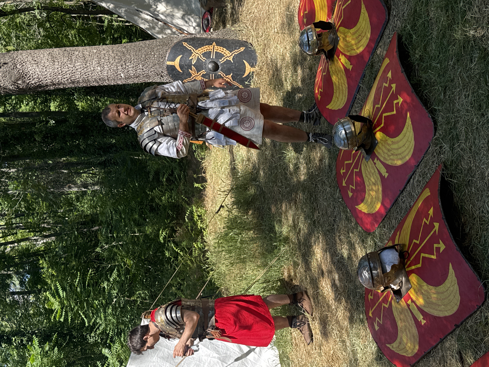The image depicts a reenactment of Roman soldiers outdoors in a wooded area. In the foreground, three Roman shields, red with yellow designs, are laid on the grass. Each shield has a Roman helmet placed on it. In the middle ground, two men dressed in Roman military attire stand. The man on the right wears a metallic segmented armor, a white tunic, and a red sash, holding a sword. He stands next to a large Roman shield leaning against a tree. The man on the left wears similar armor and a red tunic, and appears to be examining something in his hands. In the background, tents and more foliage are visible. The scene is set in the city of Plovdiv, Bulgaria, likely at an event such as the "Ancient Plovdiv" festival or a historical reenactment held in one of the city's parks or open spaces.