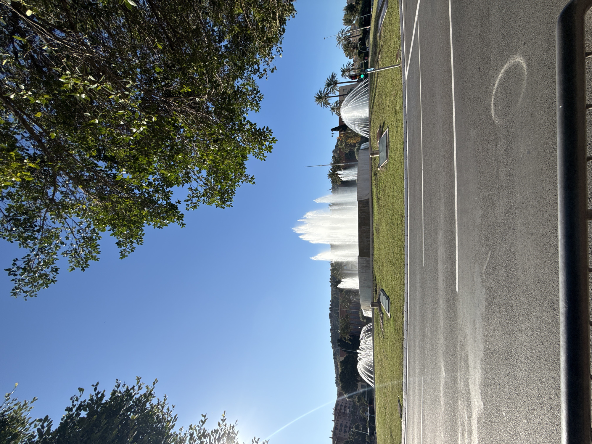 The image shows a street in an unknown city. A line of fountains are visible, spewing water upwards. The street is divided by white lines, and there is a dark pole on the right. Green foliage is visible on both sides, with palm trees among it. The sky is clear and blue.