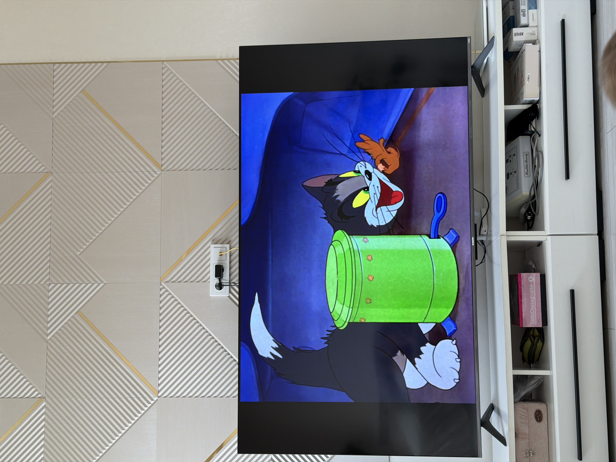 A television displaying an animated image of Tom and Jerry is the focal point of the scene. The image shows a black cat with a green object attached to its body. The television is positioned in front of a wall with a textured geometric pattern in beige tones with gold accents. An electrical outlet is visible on the wall to the left of the television. To the right of the television, a white shelving unit is present, filled with various items. The precise location in Rossiyskiy, Russia cannot be determined from the provided information.