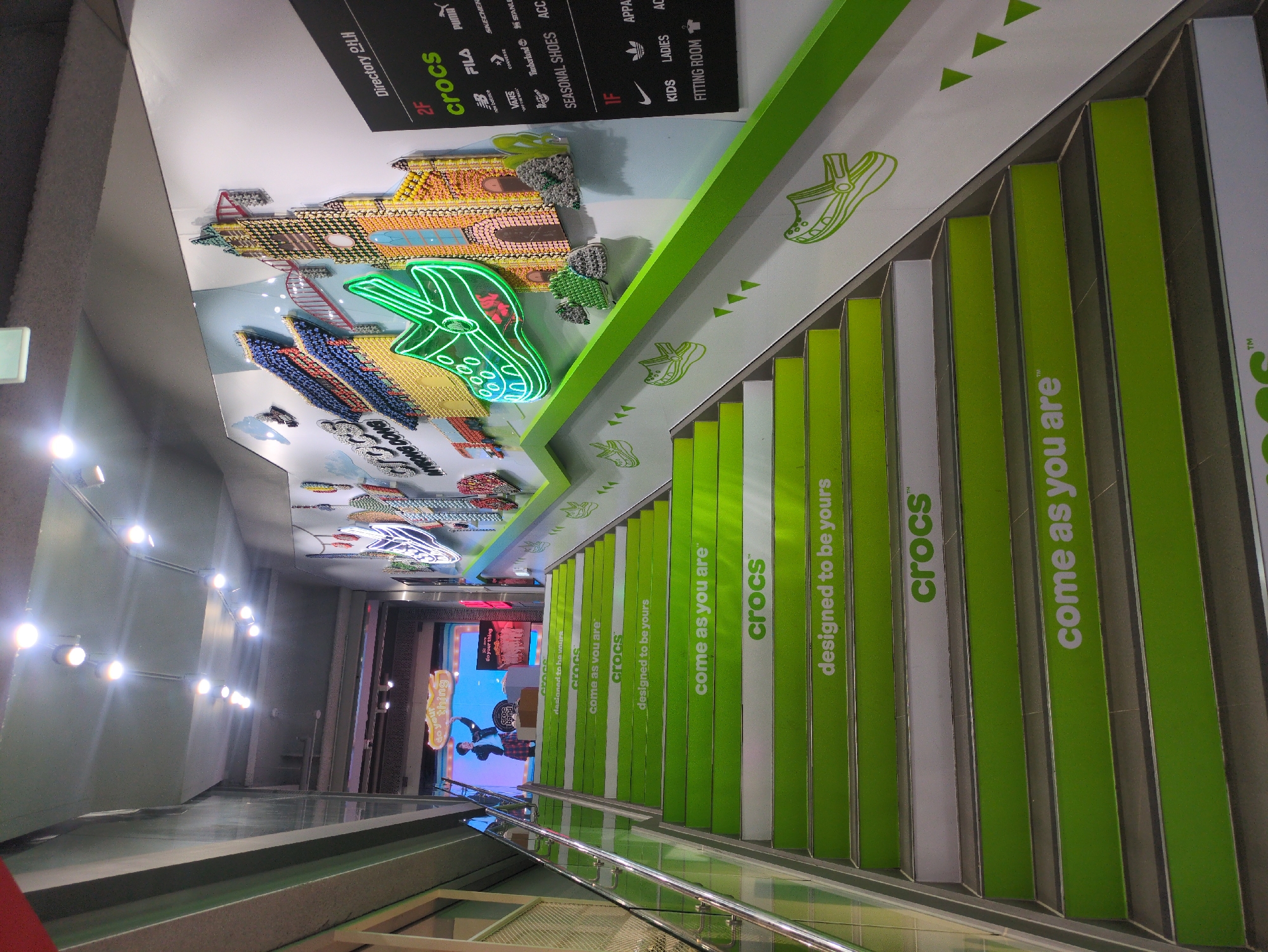 This image captures an interior view of an upward-sloping staircase within a commercial establishment, identified as a Crocs location in Myeongdong, Seoul, South Korea. The staircase consists of steps alternately colored green and white, with the green risers featuring Crocs brand slogans such as "crocs™", "come as you are™", and "designed to be yours" in white text. A transparent glass railing with a metallic top handrail ascends along the left side of the staircase.
On the right wall, a large, multicolored 3D art installation depicts various architectural structures, including a traditional Korean building and a European-style church, both seemingly constructed from small, distinct objects. A prominent neon green outline of a Crocs clog is integrated into this artwork, along with the words "crocs" and "MYEONGDONG". Adjacent to this art, a black directory board, labeled "Directory 안내", indicates "2F" (Second Floor) houses "crocs", "new balance", "FILA", "PUMA", "Vans", "Timberland", "SKECHERS", "Converse", "Dr. Martens", "Stans", "SEASONAL SHOES", and "ACC" (Accessories). "1F" (First Floor) lists "NIKE", "ADIDAS", "APPAREL", "KIDS", "LADIES", and a "FITTING ROOM". The lower section of the right wall along the staircase displays green outlines of Crocs clogs and triangular motifs.
At the top of the stairs, a large video screen shows a male individual in a plaid skirt and jacket, striking a pose or dancing, beneath a graphic bubble containing the text "do your thing". A small display stand bearing the Crocs logo and the phrase "do your thing" is positioned in front of the screen. The ceiling is grey and equipped with multiple bright, recessed spotlights illuminating the scene.