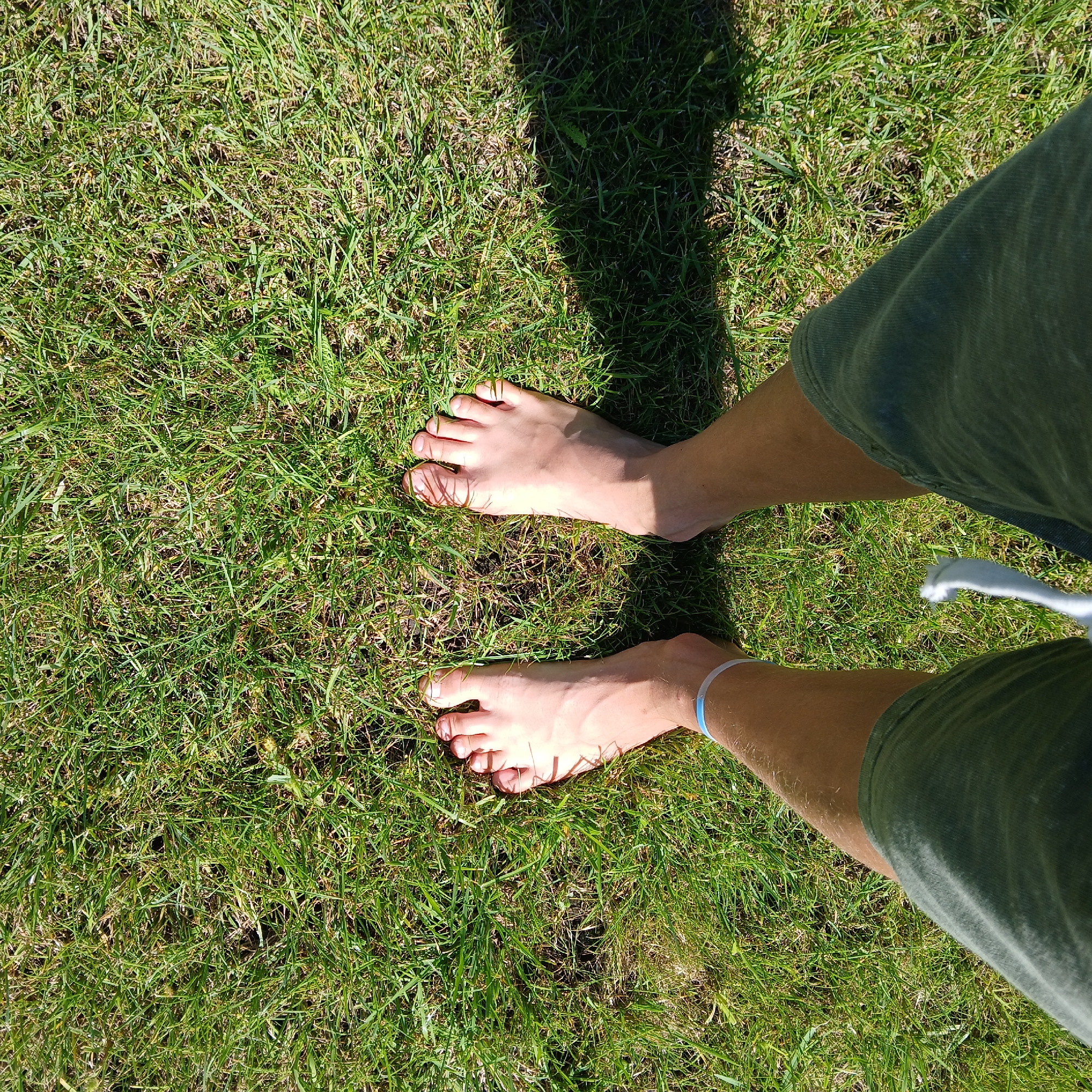 The image shows a pair of bare feet standing on green grass. The feet are positioned facing upwards, with the toes spread slightly. A blue and white bracelet is visible on the left ankle. The person is wearing dark green shorts. The grass appears well-maintained and is partially in sunlight and shadow, indicating an outdoor setting. The image does not provide any specific information that would allow for identification of the location within the city of Samarsky, Russia.