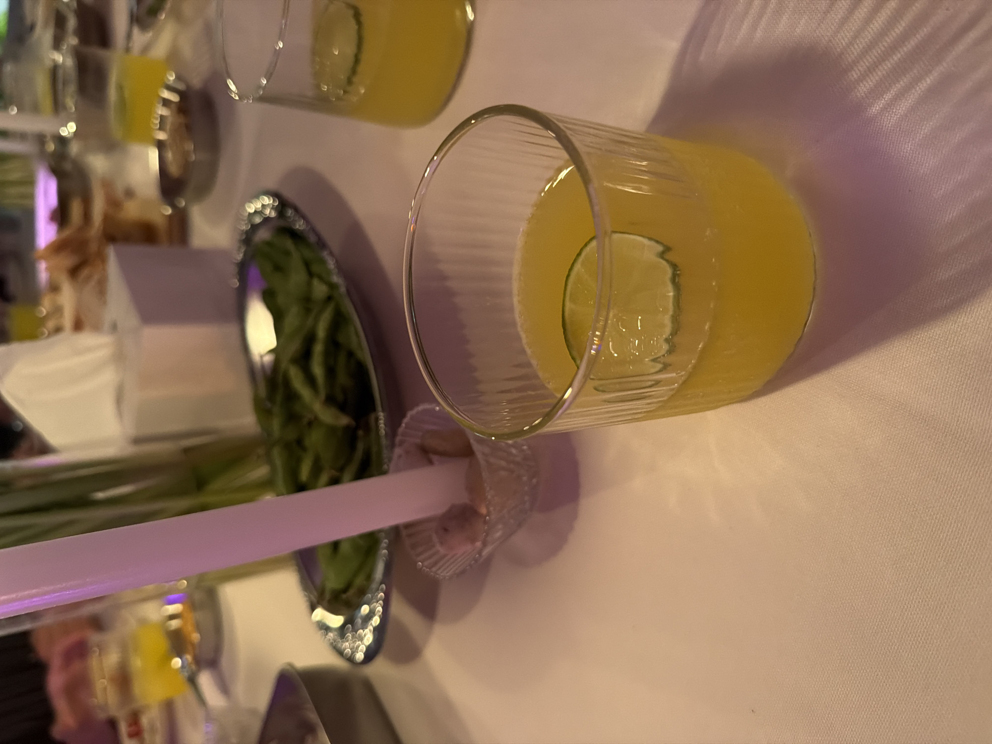 A dimly lit, formal dining scene is depicted, likely in Chang Phueak, Thailand, with a white tablecloth covering the table. In the foreground, a clear glass filled with a bright yellow beverage and a slice of lime is prominently featured, its textured surface catching the ambient light. To the right, another glass of the same yellow drink is partially visible. In the mid-ground, a silver serving dish holds a pile of green edamame, and a white candle stands upright in a small, crystal dish containing a light-colored substance, possibly salt. Behind the edamame, a white tissue box and other food items on silver platters are scattered across the table. The lighting is soft and diffused, with hints of purple and pink hues, creating a warm and intimate atmosphere.