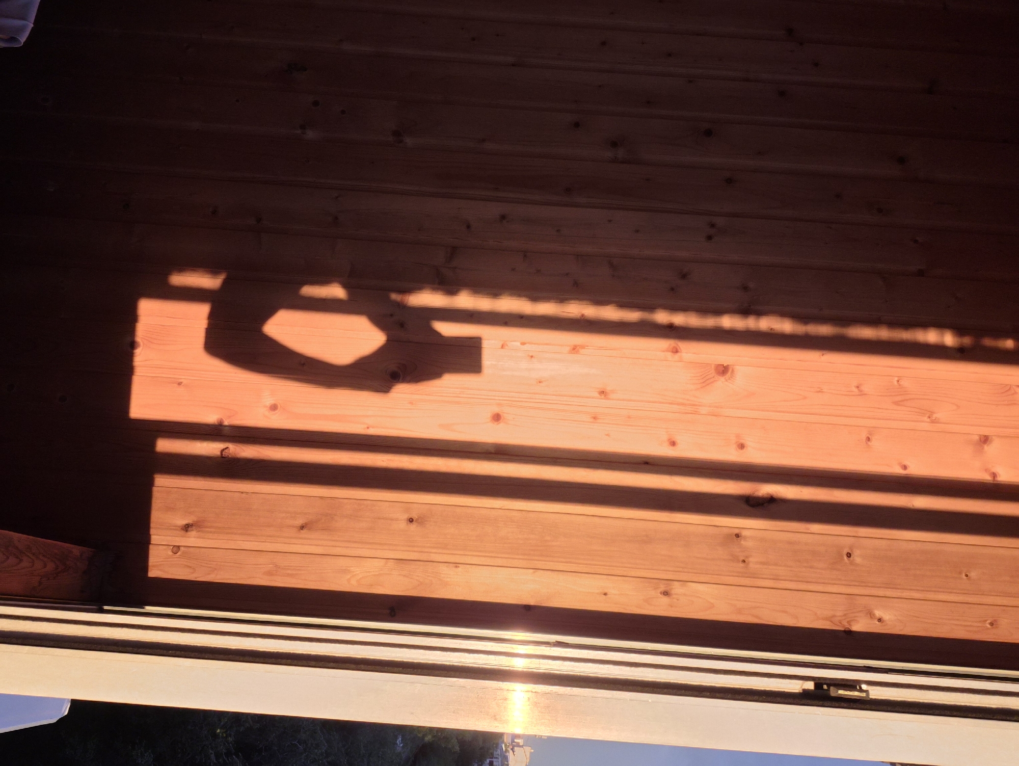 The image displays a close-up view of a wooden wall bathed in the warm, orange glow of late afternoon sunlight. The wood grain is clearly visible, with vertical planks and scattered knots. A distinct shadow, cast by what appears to be a person holding a camera or phone, is prominently featured on the left side of the wall. The shadow shows a head, an arm extended to hold a device, and the shape of the device itself. Parallel shadows from vertical elements, possibly a fence or window frame, stretch across the wall from the right. The right edge of the frame reveals a sliver of the outdoors, with a hint of a building and greenery visible in the distance, illuminated by the setting sun. The scene is devoid of any specific landmarks that would definitively place it in Chita, Russia; it is a generic outdoor setting.
