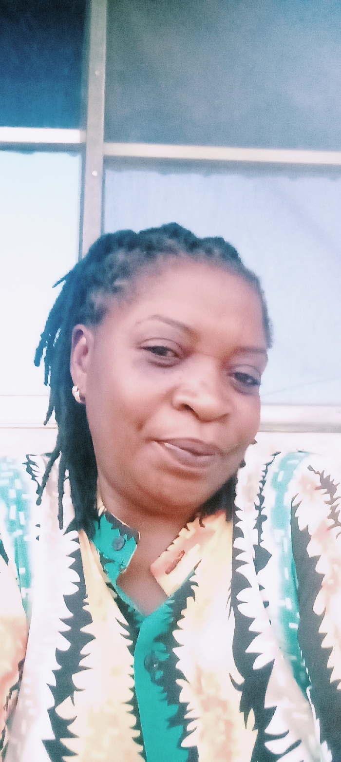 A woman with dark skin, braided hair, and gold hoop earrings smiles slightly. She wears a colorful button-down shirt with a repeating pattern of black and white "leaf" shapes on a yellow and light orange background, with teal vertical stripes on the button placket. Behind her, a window with a grid pattern is visible, with varying shades of blue and white. The overall image appears to be taken indoors under natural light.