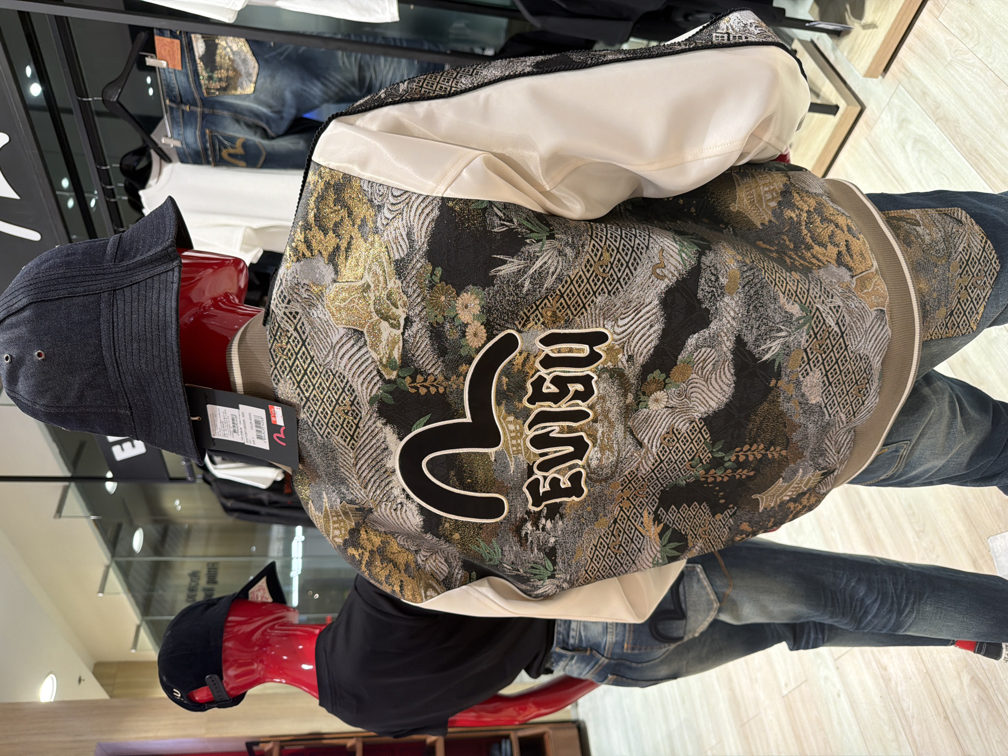 This is a full-shot image, taken at eye-level, of two mannequins in a retail store. The mannequin on the right is wearing a black bucket hat, a jacket with a cream-colored satin lining and a complex, ornate Japanese-style print on the back, and blue jeans. The print on the back of the jacket features a landscape scene with mountains, trees, and a building, all rendered in a textured pattern of gold, black, white, and green threads. The Evisu logo is prominently displayed in black outline on the upper back of the jacket, with the brand name "EVISU" written below it in gold embroidery. The mannequin on the left is wearing a red baseball cap, a black t-shirt, and blue jeans. The jeans have the Evisu logo embroidered in black on the back pocket. The background shows a retail environment with clothing racks holding various garments, including white t-shirts and denim jeans. The lighting is bright and even, suggesting an indoor commercial setting. The floor is a light-colored wood. There are no identifiable people in the image; the subjects are mannequins. The location is a retail store in Ko Samui District, Thailand.