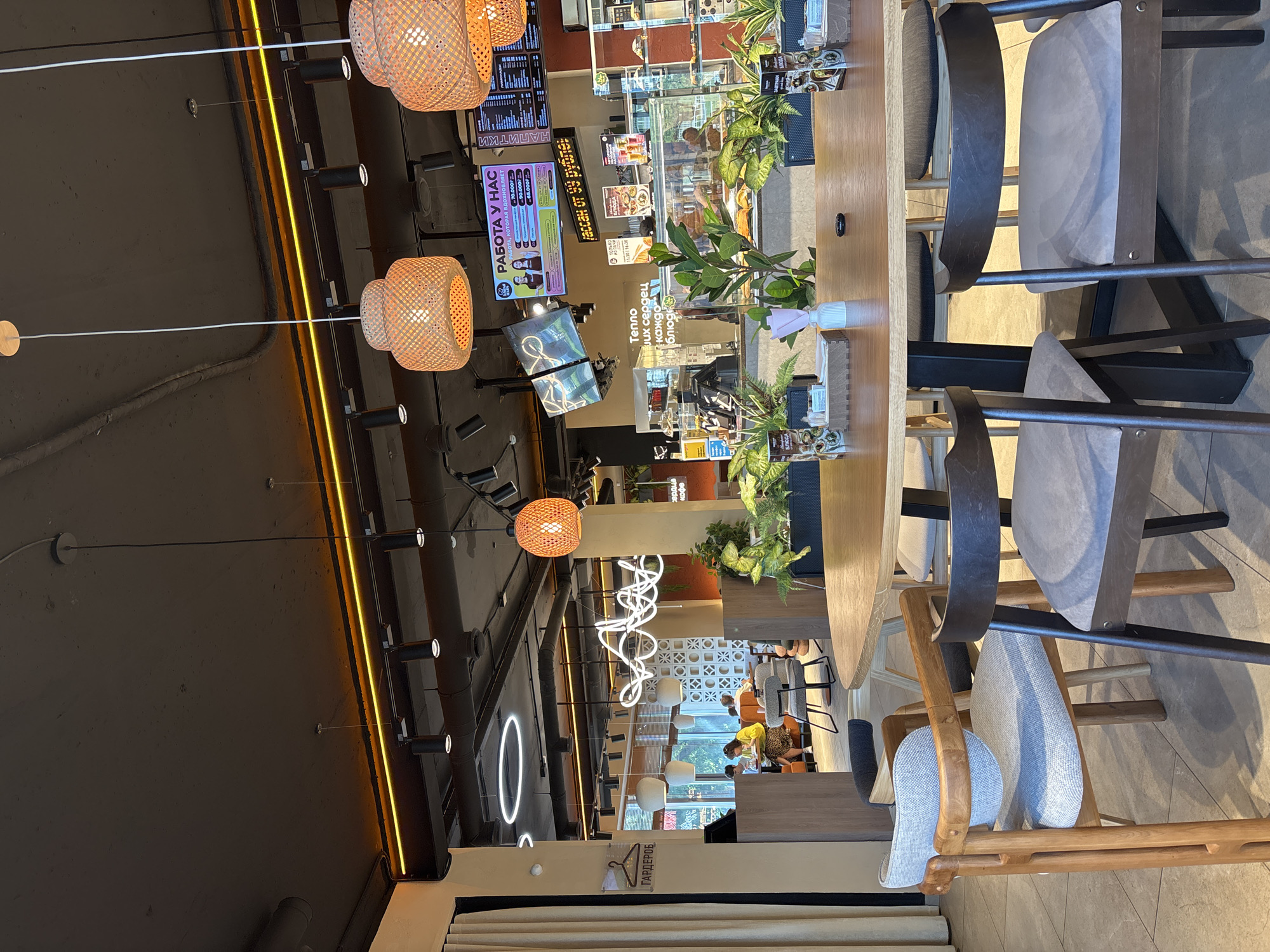 The image shows the interior of a modern cafe. In the foreground, a large wooden oval table is set with several chairs around it. The chairs have wooden frames and upholstered grey seats and backs. One chair, in the foreground left, has a lighter wood frame and a lighter grey fabric seat. Another chair to its right has a darker wood frame and a similar grey seat. Further back, more chairs are visible around the table and other seating areas.

In the middle ground, a counter area is visible with food displays and a cashier. Plants are placed around the counter and in the seating area, adding a touch of greenery. Overhead, decorative pendant lights with woven shades hang from the ceiling. The ceiling itself is dark and industrial-looking, with exposed pipes and lighting fixtures. A large screen displaying job advertisements is mounted on the wall above the counter.

In the background, through a window or open area, other patrons can be seen seated at tables. The cafe appears to be well-lit, with a mix of ambient and task lighting. The overall atmosphere is casual and contemporary. There is no specific visual information in the image that directly indicates the city of Naberezhnye Chelny, Russia.