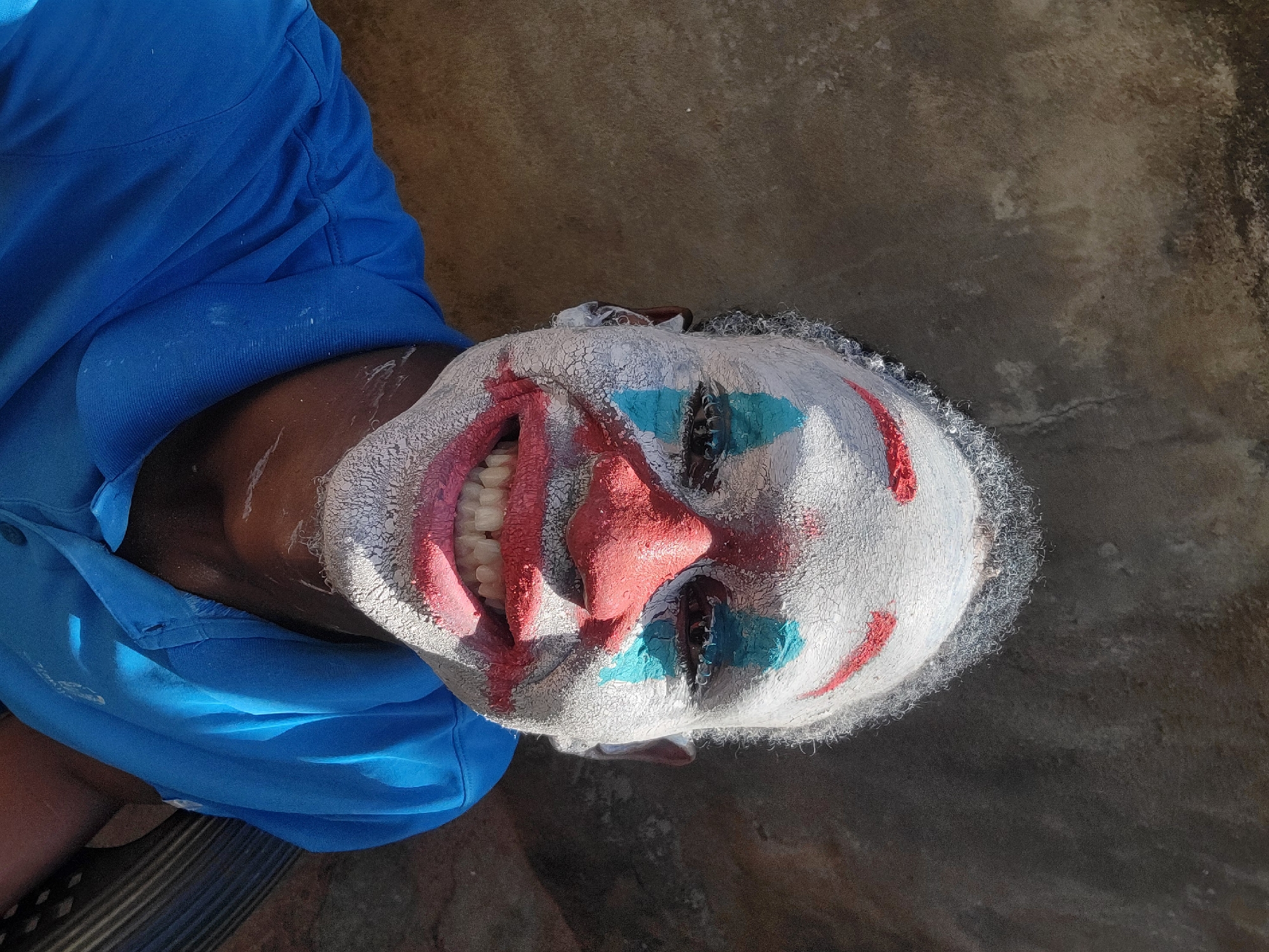 A person is lying on a concrete surface in an outdoor setting, with the head oriented towards the top of the frame. The individual is wearing a white clown mask with painted red details around the mouth and on the nose, and blue markings around the eyes. The mask appears to be cracked and textured. The person is wearing a blue shirt. The setting appears to be in Nkpor, Nigeria, as specified in the prompt.