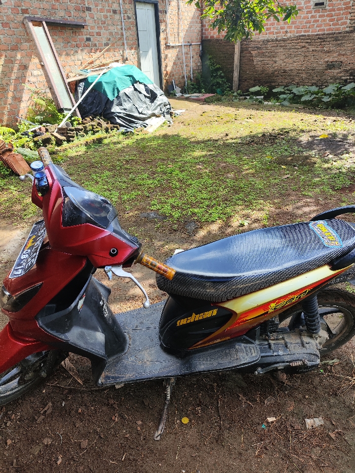 An outdoor, eye-level shot captures a red and black scooter parked on a dirt ground with sparse green vegetation, likely in a backyard setting in Pubian, Indonesia. The time of day appears to be sunny daytime, casting subtle shadows.

The scooter, positioned slightly angled towards the right, occupies the foreground and mid-ground. Its front fairing is a metallic red, while the footrest area is black. The seat is covered with a textured, possibly carbon-fiber-patterned material, and the side body panel features yellow and orange graphics. Below the seat, text resembling "oohan" is visible. The scooter's kickstand is down, suggesting it is parked. A partial license plate is visible on the front fender, showing "41.86" and some indistinguishable letters.

In the background, a brick building wall stands prominently, indicating an older structure. An unpainted white door is set into the wall on the left. To the left of the door, leaning against the wall, are what appear to be old window frames or pieces of wood. Further left, a pile of miscellaneous items covered with green and black tarps suggests storage or discarded materials. The ground in the mid-ground and background consists of patchy grass and dirt, with some larger green foliage visible on the far right. No people or specific activities are depicted.