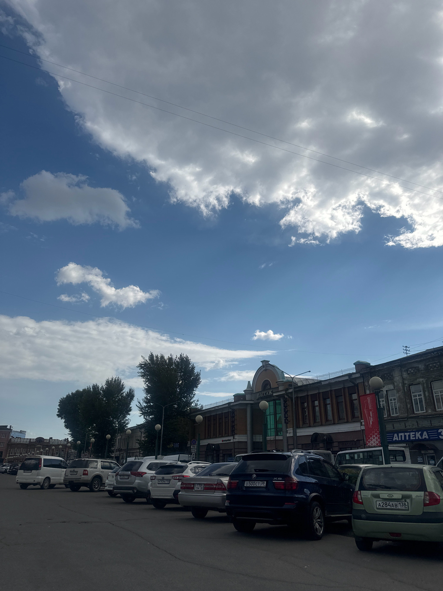 The image depicts a street scene in Irkutsk, Russia, on a partly cloudy day. In the foreground, a row of parked cars lines the asphalt road. These vehicles include a dark blue BMW X5, a light green hatchback, and several other sedans and SUVs. In the background, a long, multi-story building with a historic facade runs along the street. The building features arched windows and signage, with one sign reading "АПТЕКА 3" (Apteka 3, meaning Pharmacy 3) and another "ПАССАЖ" (Passazh, meaning Arcade). Trees with dark green foliage are visible to the left of the building. The sky above is a mix of blue and grey clouds, with power lines stretching across the upper portion of the frame. There are no people clearly visible in the image.