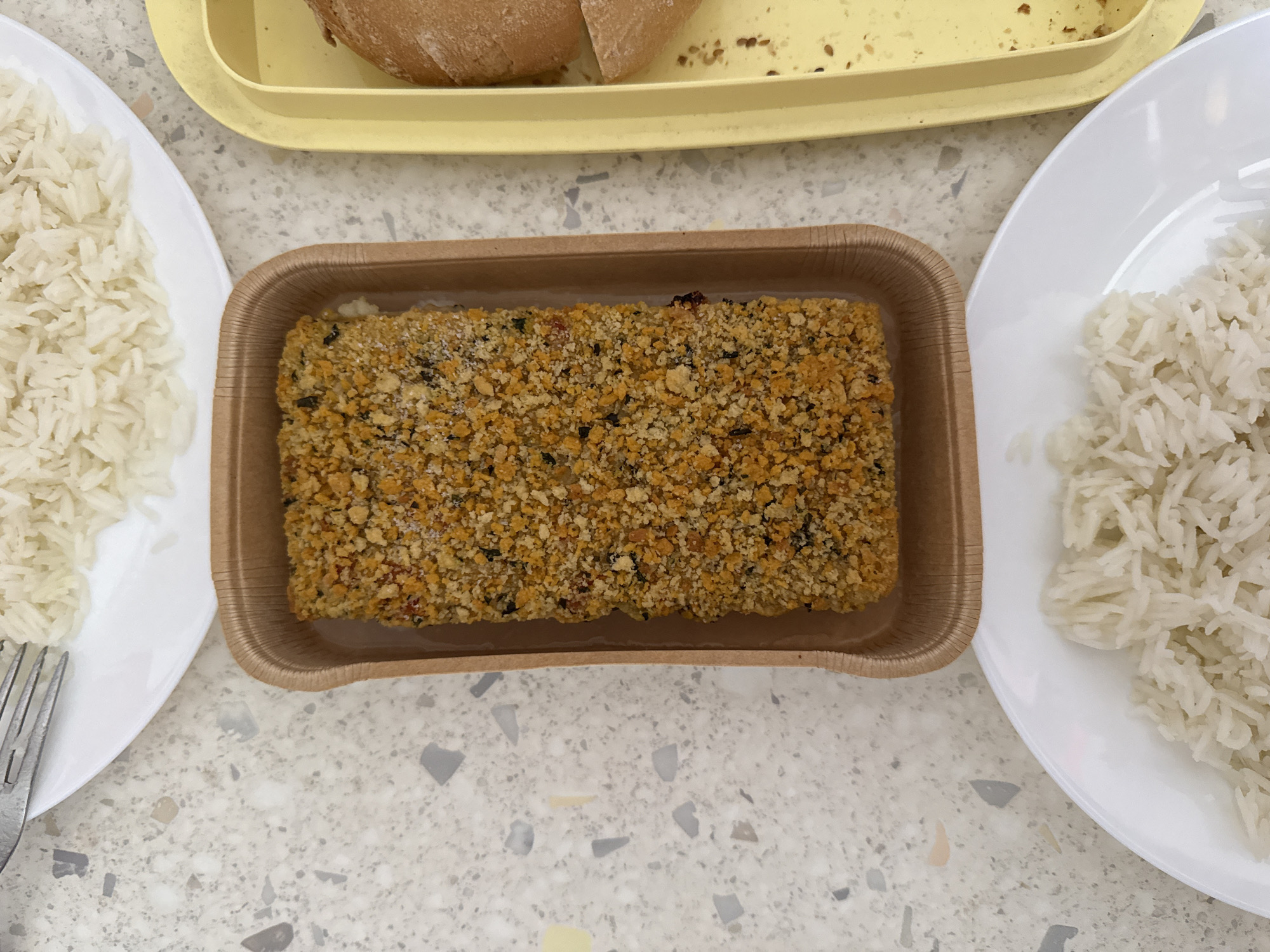 A top-down view shows a meal laid out on a speckled grey countertop, likely in Milan, Italy, as suggested by the context of the image. In the center is a rectangular brown paper tray holding a breaded food item, possibly chicken or fish, covered in coarse golden-brown breadcrumbs with flecks of green herbs and red spices. To the left of the tray, a portion of white rice is visible on a white plate, next to a metal fork. To the right of the tray is a yellow plastic tray containing two halves of a bread roll. Another white plate with white rice is positioned in the lower part of the image. The lighting is bright and even, suggesting an indoor setting. No people are visible in the image, and there are no specific architectural features to identify the exact location within Milan. The overall scene depicts a simple, prepared meal.