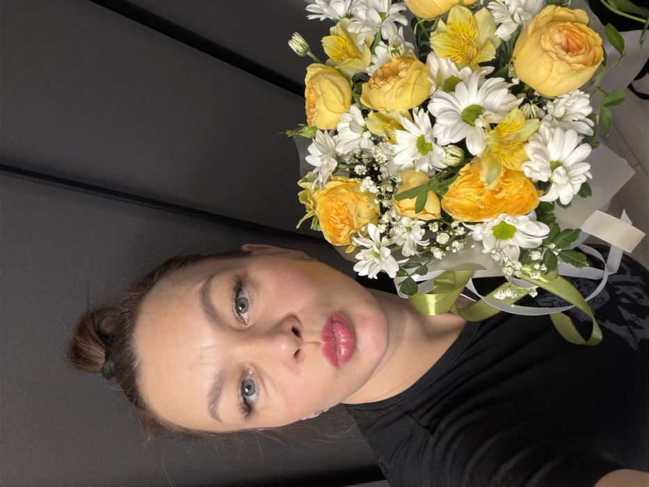 The image displays an adult female in a selfie-style photograph, situated in the city of Chaikovsky, Russia. She has brown hair pulled into a top bun and light-colored eyes, gazing directly at the camera. Her lips are pursed in a distinct gesture. She is wearing a black short-sleeved t-shirt, with her right shoulder and arm visible on the left side of the frame. She holds a round bouquet of flowers in the lower right portion of the image. The bouquet consists primarily of yellow roses, white daisy-like flowers, yellow alstroemeria, small white filler flowers, and green foliage. It is wrapped in a clear or translucent material, secured with light green and white ribbons. The background is a dark, solid grey wall, featuring subtle diagonal lines in the upper right quadrant.