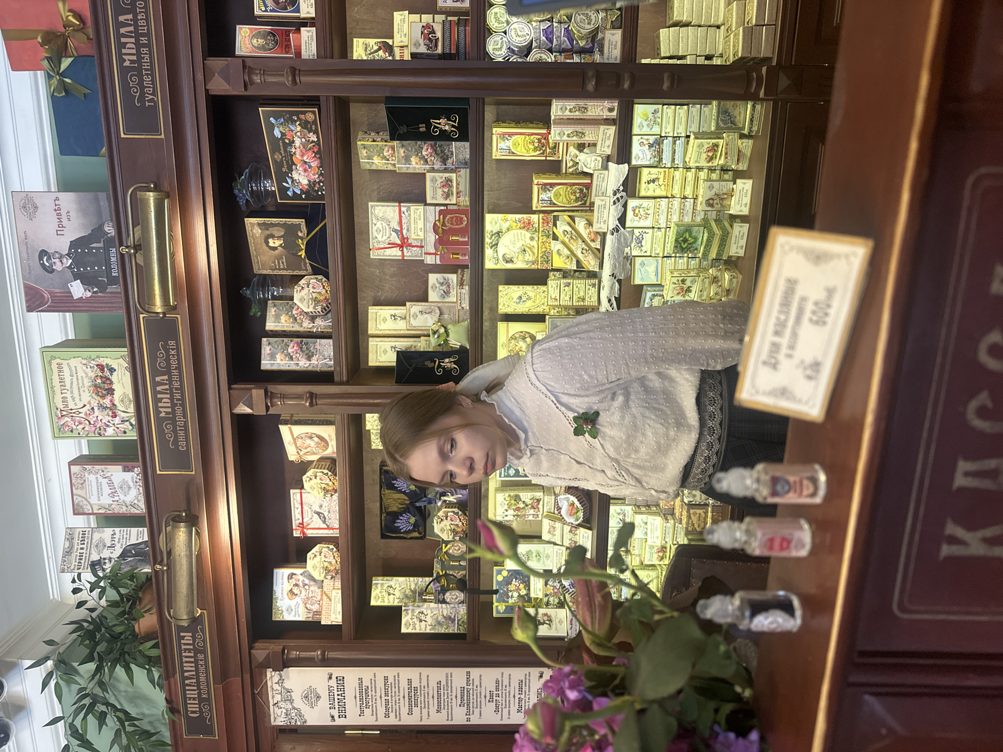 The image depicts the interior of a vintage-style shop in Kolomna, Russia. The shop appears to be a perfumery or soap shop, with shelves stocked with numerous boxes and bottles of what appear to be soaps, perfumes, and other cosmetic products. The products are displayed in ornate, often floral-themed packaging, suggesting a historical or retro aesthetic.
In the foreground, a young woman with light brown hair is standing behind a counter. She is wearing a white blouse with a ruffled collar and a dark skirt. She is looking to her right, seemingly at the products on the shelves. On the counter in front of her are three small glass bottles with clear liquid inside, and a sign with Russian text and pricing information.
The shelves are made of dark wood and are filled with a variety of items. There are boxes with illustrations of flowers, people, and historical scenes. Some boxes are stacked, while others are individually placed. The shop's interior design features wooden shelving, dark wood paneling, and brass fixtures, reinforcing the historical ambiance. Signs in Russian are visible on the shelves and walls, some with decorative lettering. A hanging sign with Russian text is visible on the left side of the image. The lighting is warm and seems to come from overhead fixtures and possibly from within the display cases. The overall impression is of a well-preserved historical establishment selling traditional products.