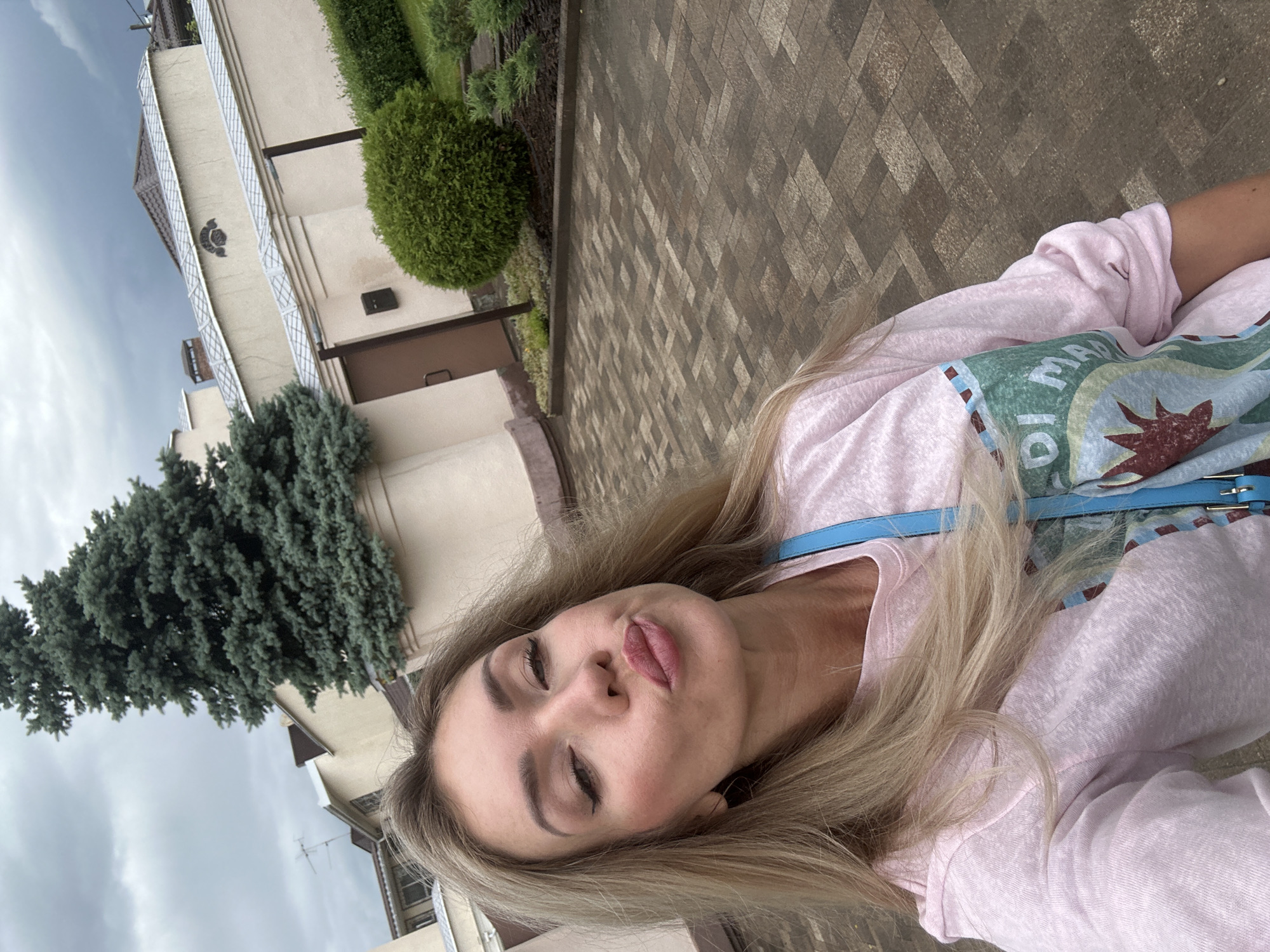 A woman with blonde hair and light skin is taking a selfie. She is wearing a pink sweatshirt with a graphic design on it and a blue strap across her chest. Her lips are pursed, and she is looking directly at the camera. The background shows a paved walkway, a tall evergreen tree, a building with a cream-colored facade, and a manicured garden. The sky is overcast. The image appears to be taken outdoors in a residential or institutional setting.