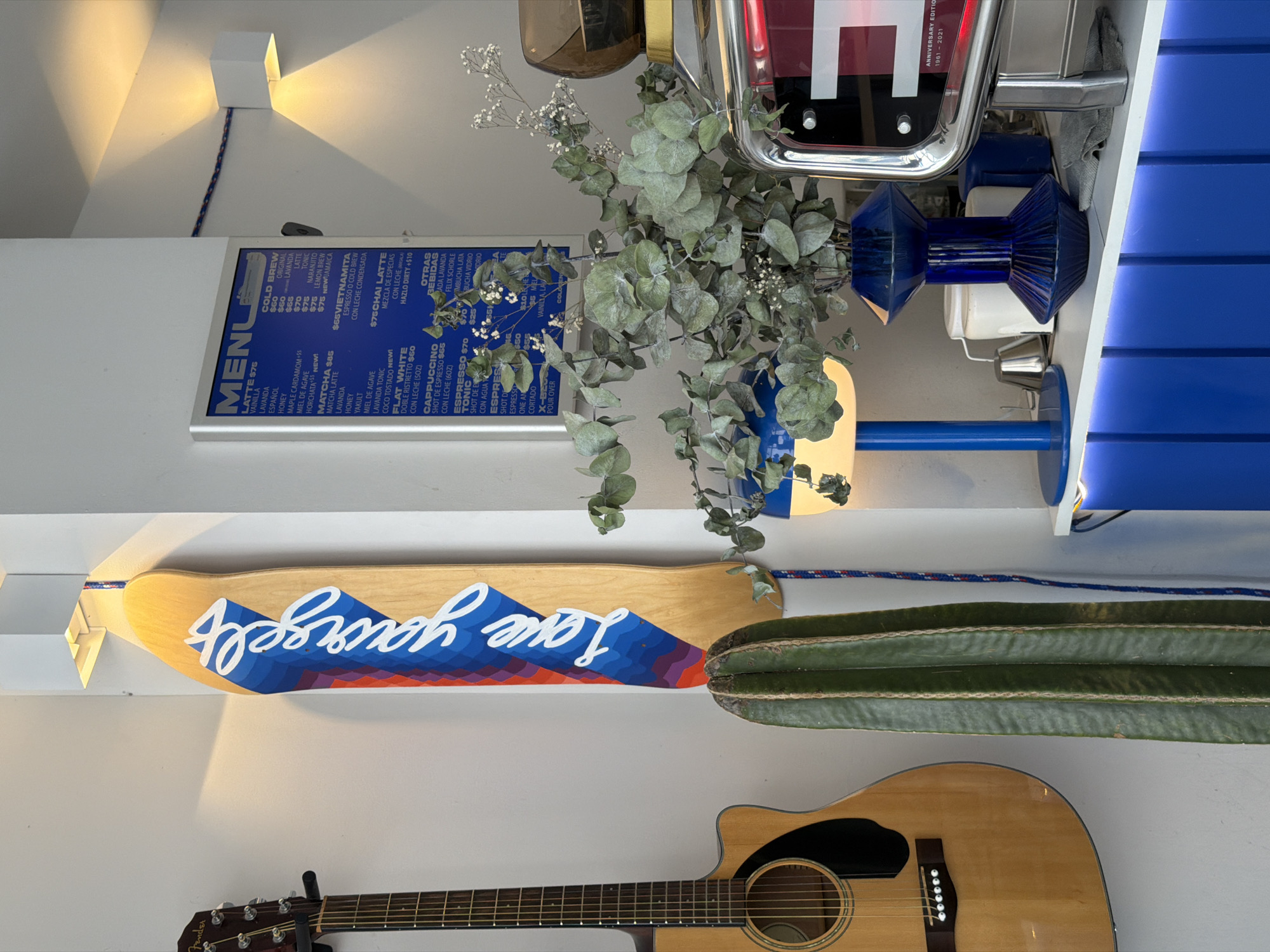 A vertically oriented indoor shot depicts a wall adorned with various items. Starting from the top, a square light fixture emits a warm glow. Below it, a blue-toned menu board lists coffee variations and prices, followed by a bouquet of eucalyptus. Adjacent to the bouquet, a vintage-style object hangs.
Further down, a skateboard with the inscription "Love yourself" is mounted above a cactus. An acoustic guitar rests below the cactus. The wall background is a plain light color. The scene takes place in San Bernardino Tlaxcalancingo, Mexico.