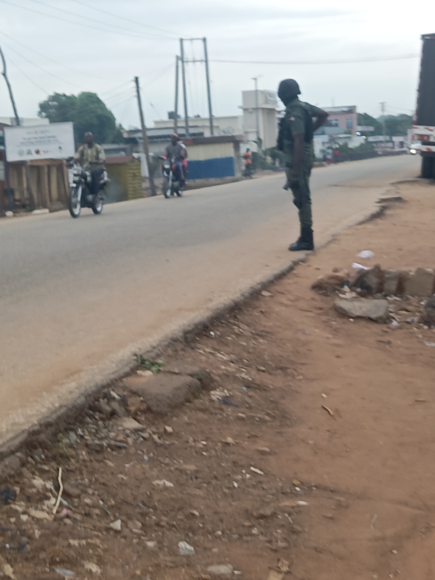 Akwanga, Nigeria, 2023. A soldier in green fatigues stands on a curb along a paved road with two motorcyclists visible in the background. Debris litters the ground.